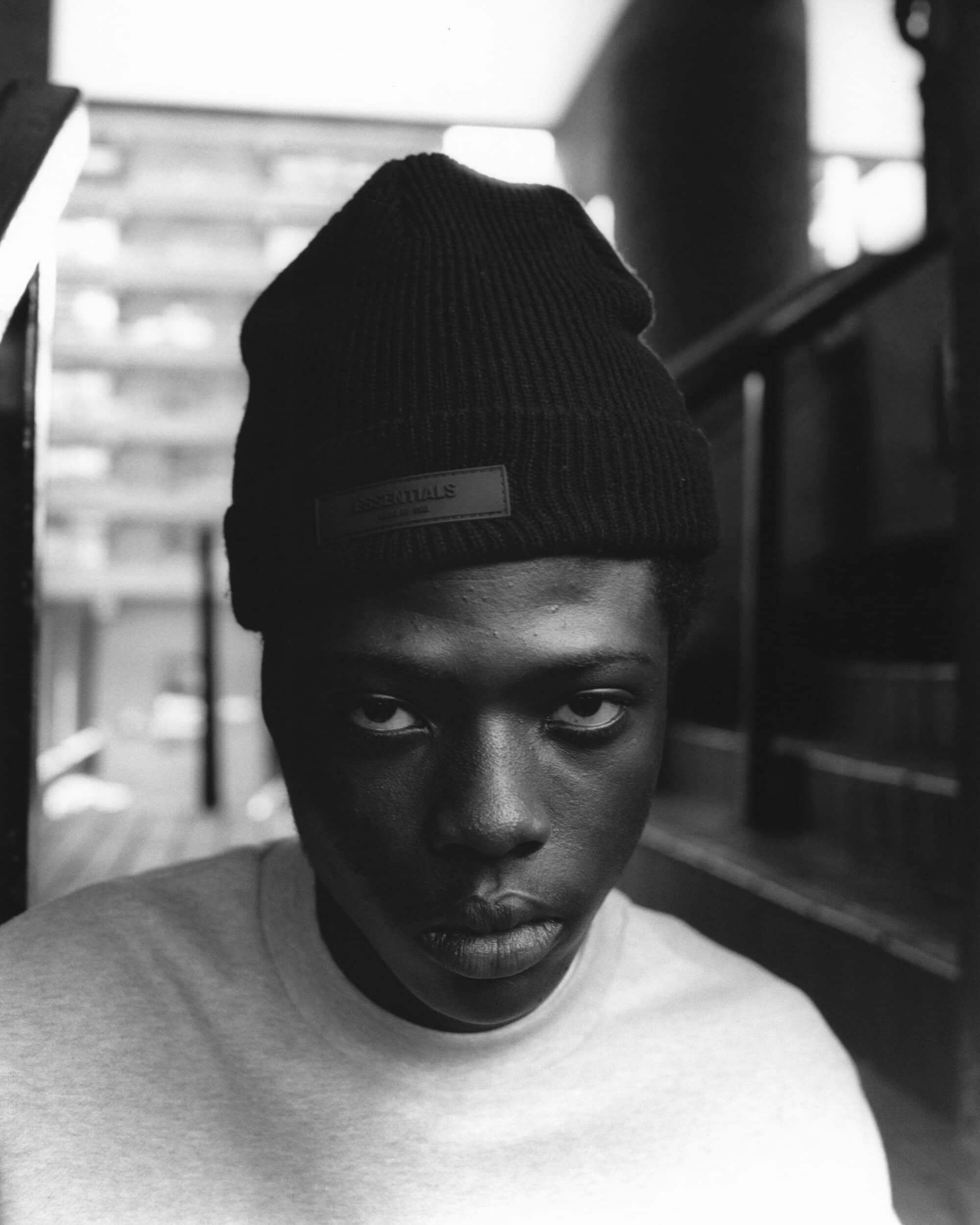 Portrait of a black man wearing a dark beanie and light gray shirt, captured in a monochrome photograph against a stairwell backdrop.