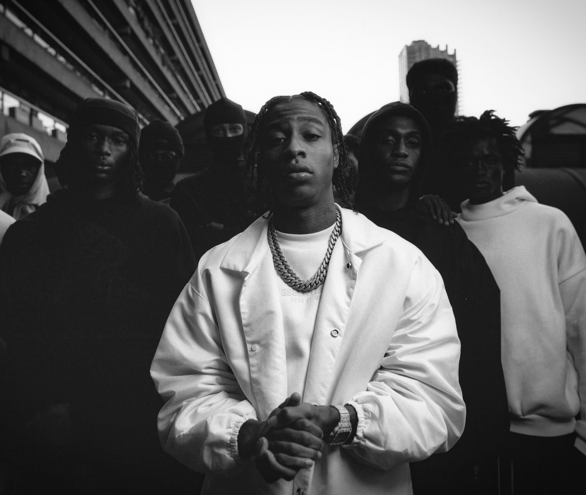 Black and white portrait of a young man in a white jacket, posing in front of a group of people and a building in an outdoor setting.