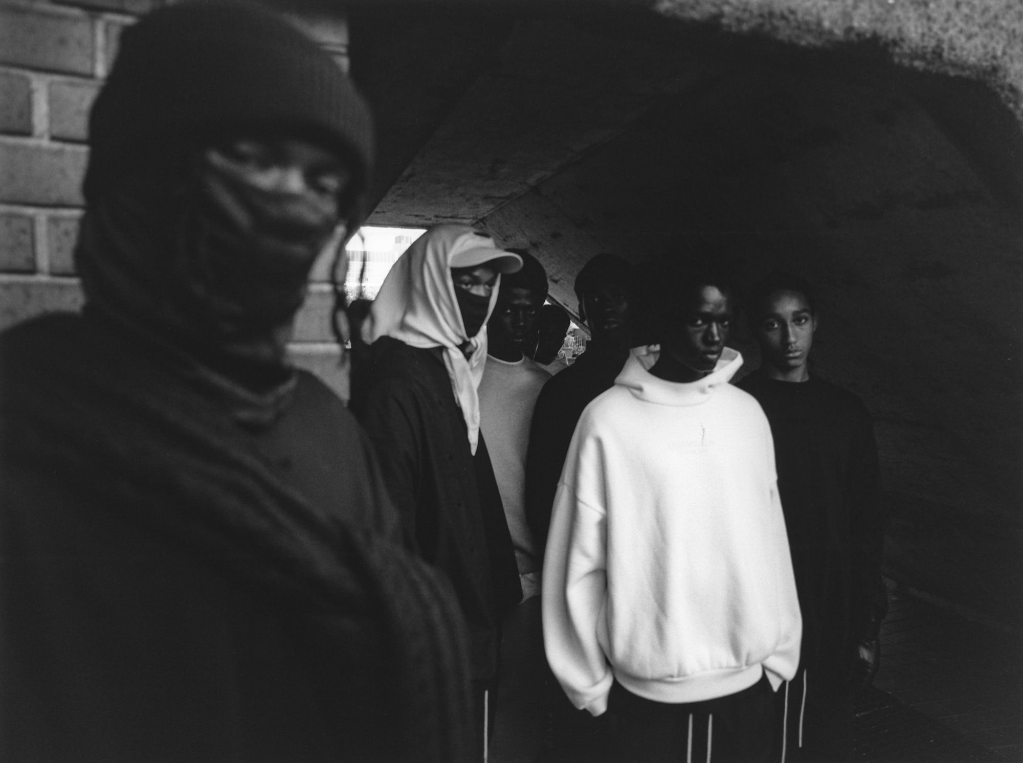 Black and white photograph of a group of young African men standing in an urban underpass, some wearing hoodies and face coverings.