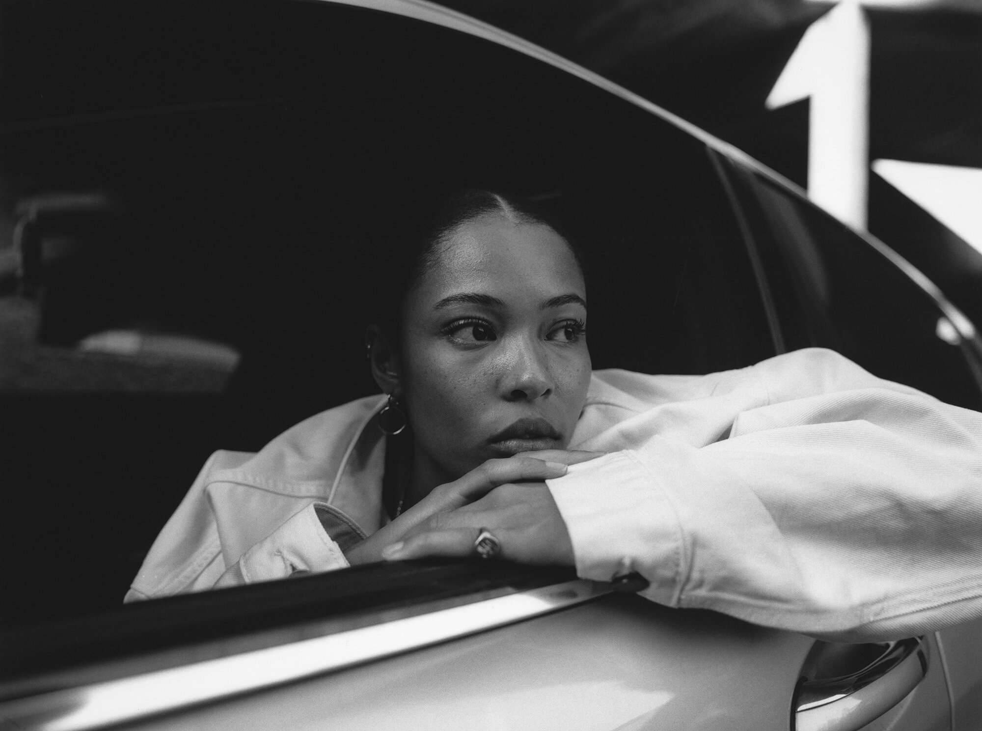Black and white shot of an African-American woman in a car with a denim jacket. She rests her chin on her hands.