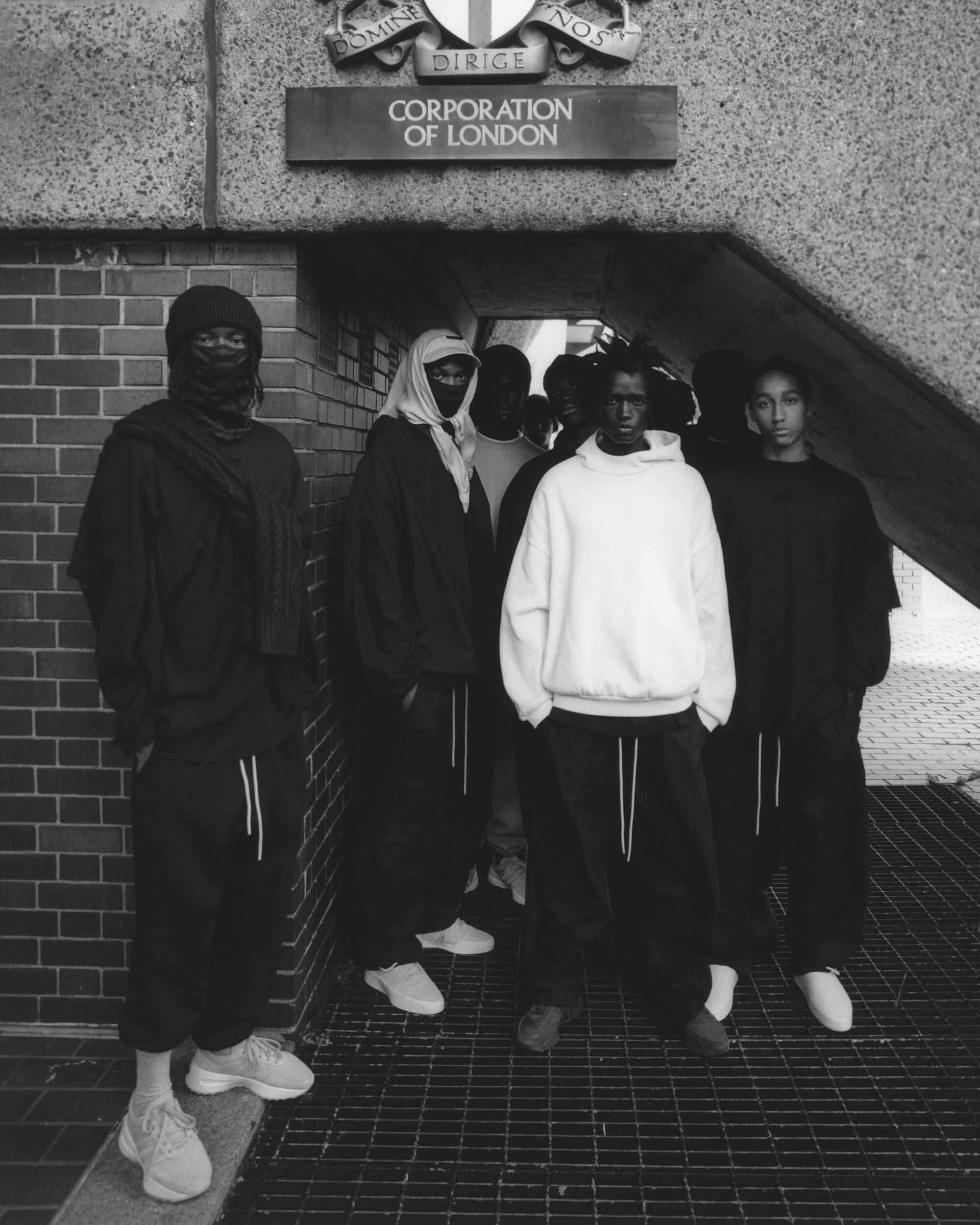 Group of men and women in streetwear fashion standing beneath the Corporation of London sign; black and white photography of urban scene.