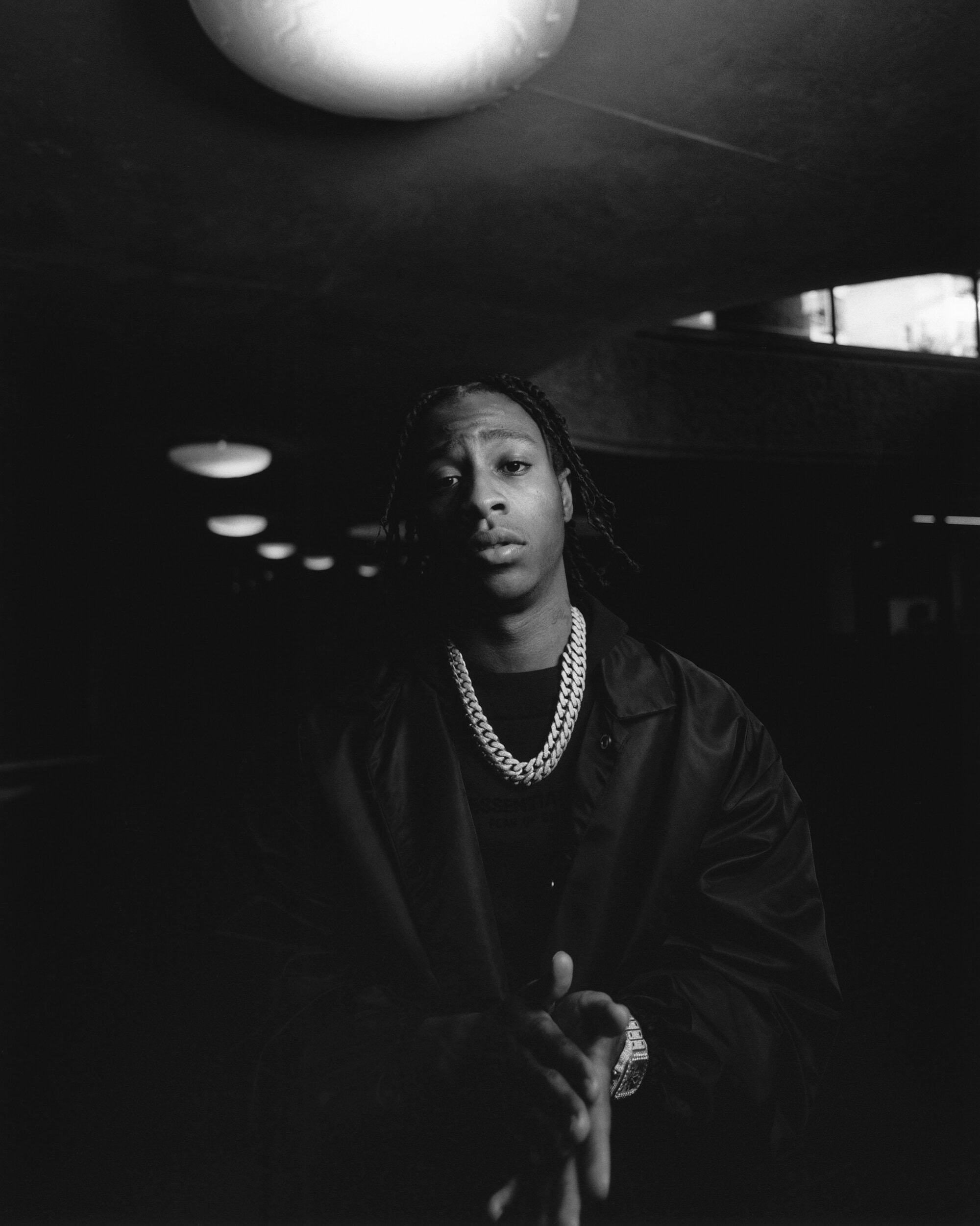 Monochrome portrait of a young adult with dark hair, wearing a chain necklace and jacket, illuminated by overhead lights in a dark space.