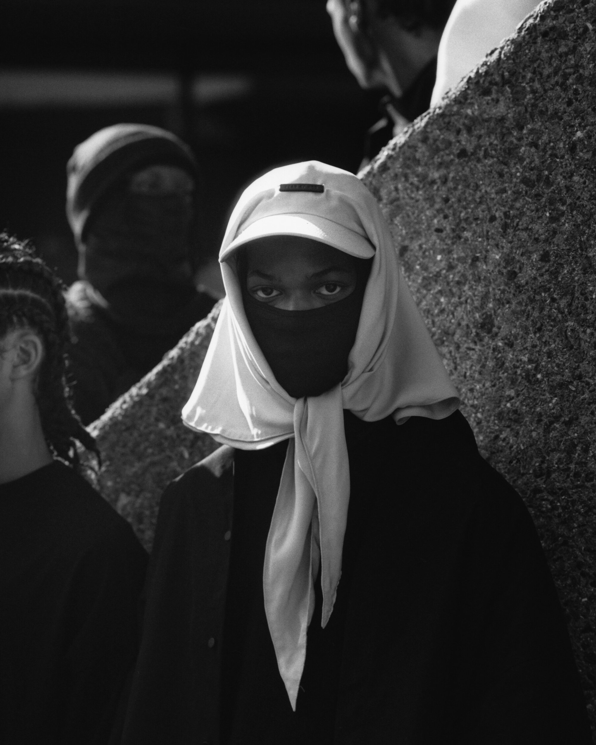 Monochrome portrait of a person with a face covering and draped headscarf standing in front of a rough concrete structure.