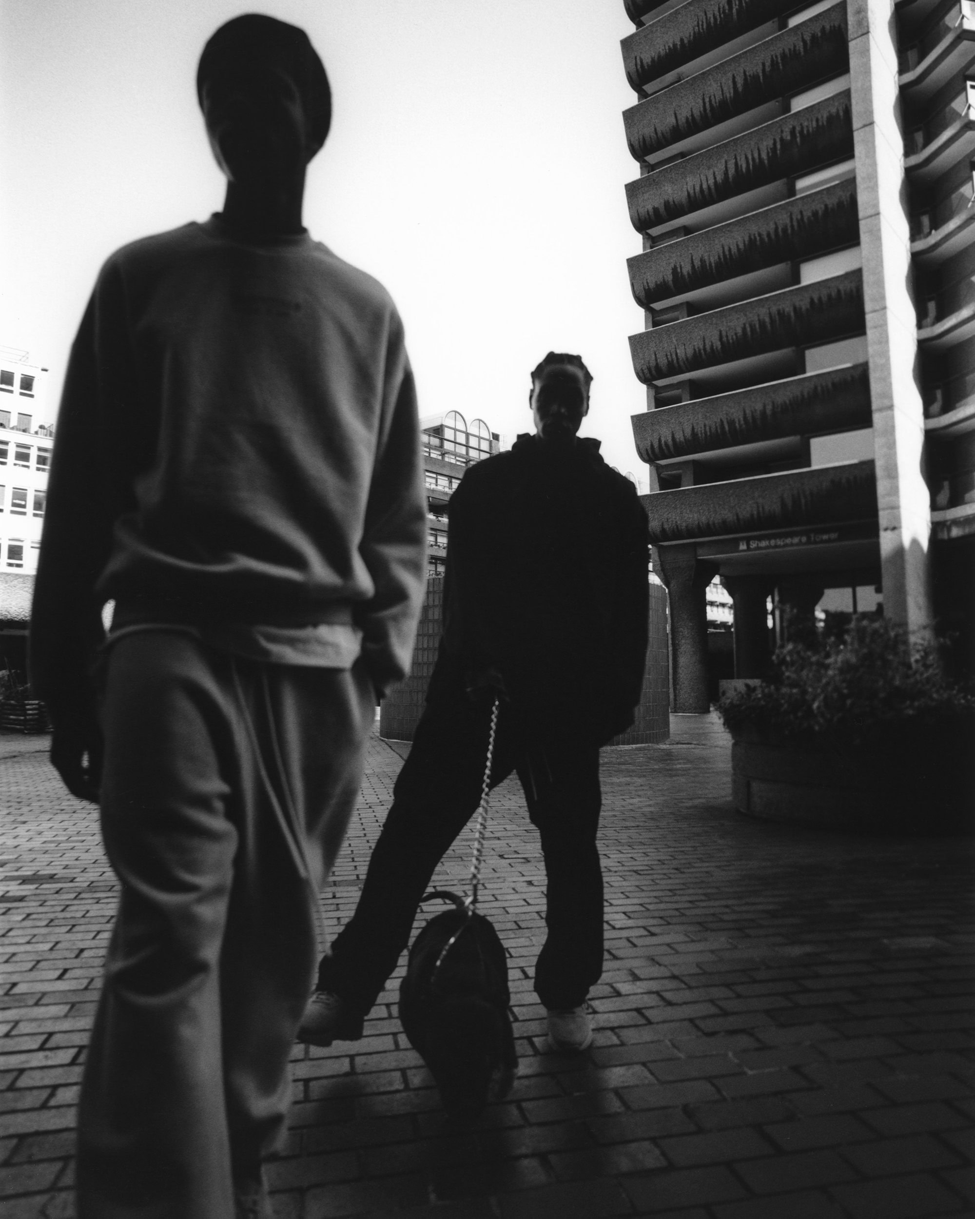 Two people stand on a brick walkway near a tall, brutalist building in an urban setting, rendered in black and white with muted tones.