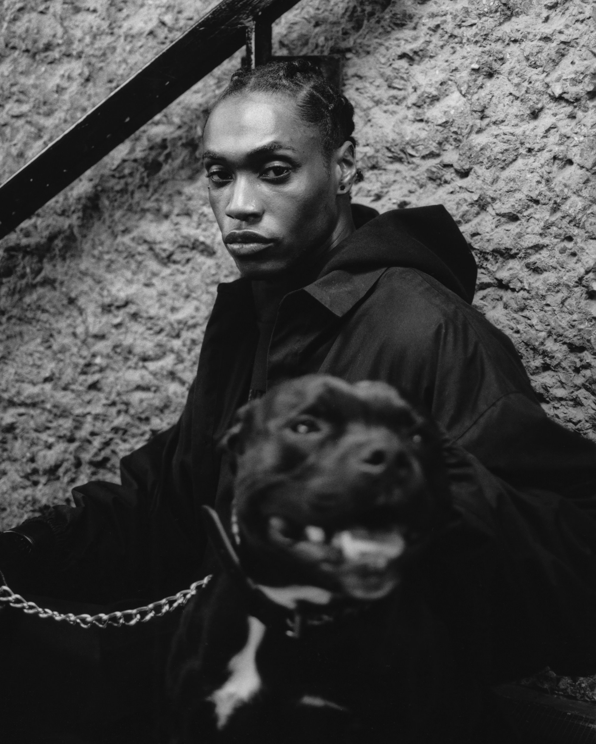 Monochrome portrait of a dark-skinned person with braids and a black hooded jacket, posing with a black dog on a chain.