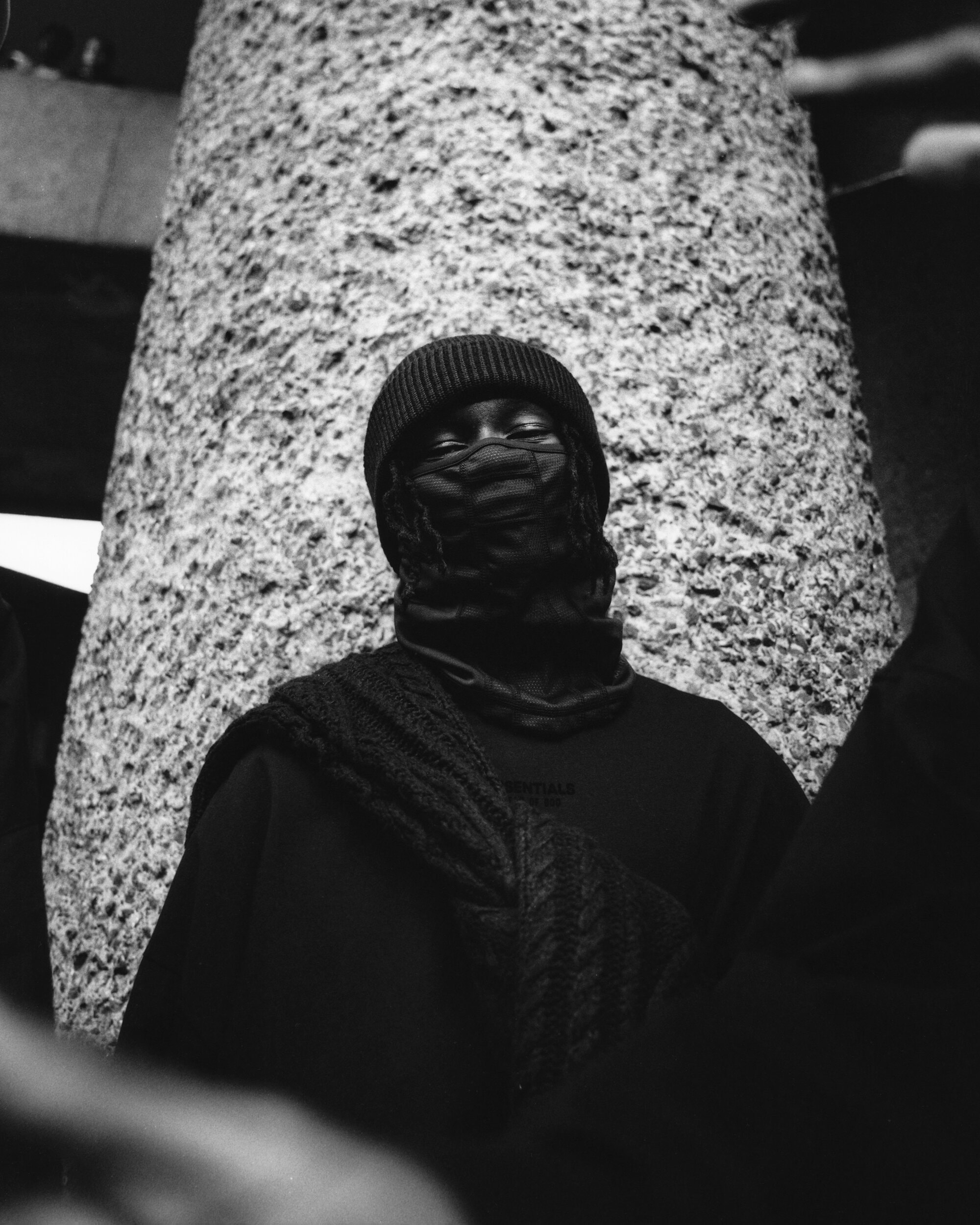 Monochrome portrait of a person in a knit hat and scarf standing against a textured wall in an outdoor environment.