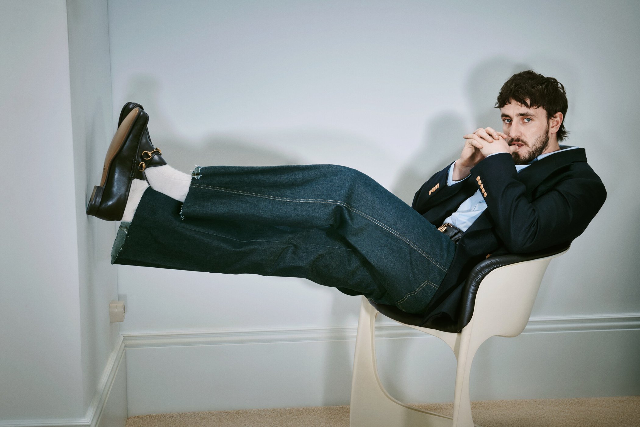 Adult male seated wearing a dark blazer, light blue shirt, wide-leg denim jeans, white socks, and loafers with gold hardware in a minimalist room.