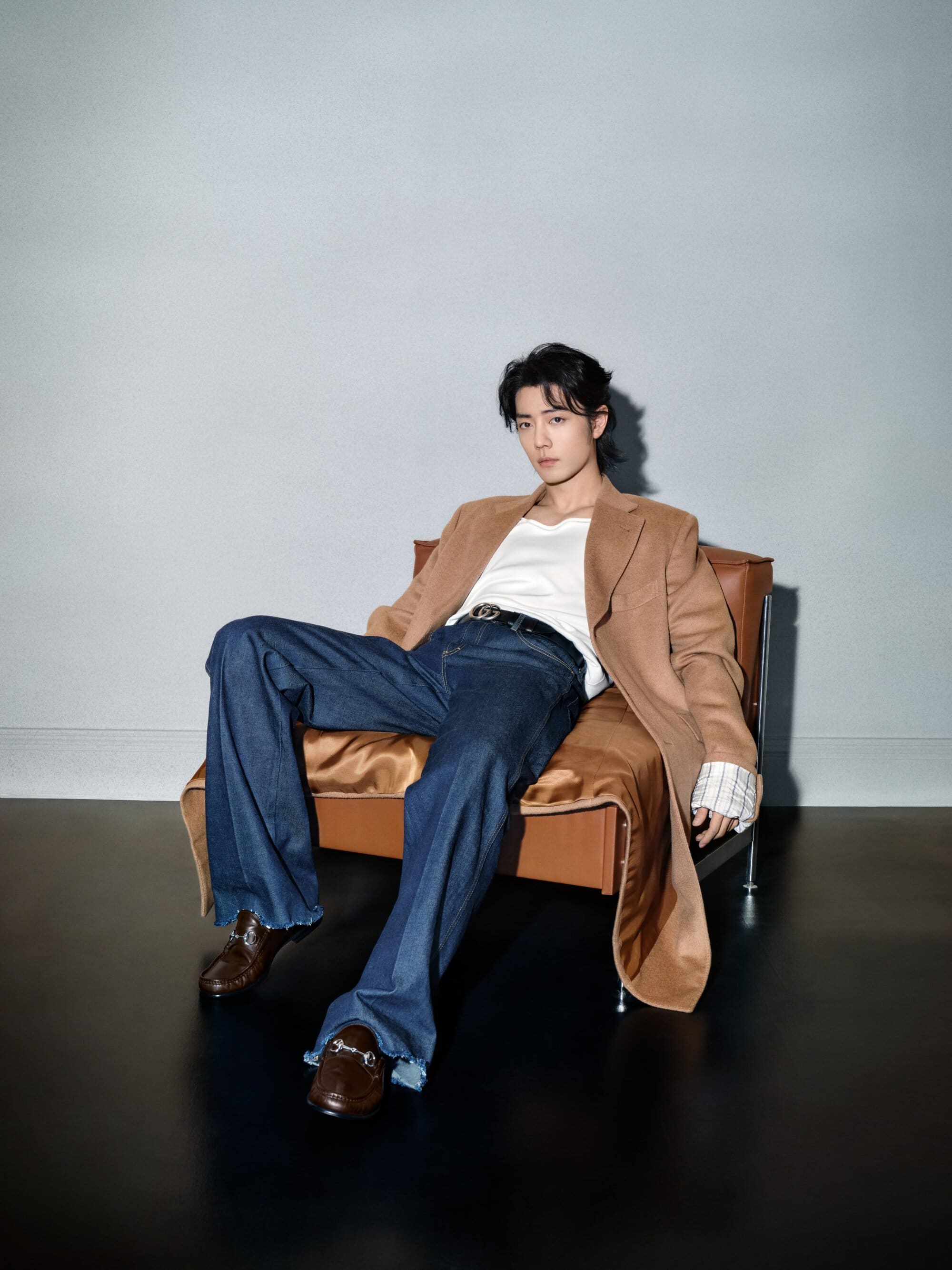 An Asian male model lounges in a brown leather chair, wearing a tan jacket, white t-shirt, dark blue jeans, and brown loafers against a grey backdrop.