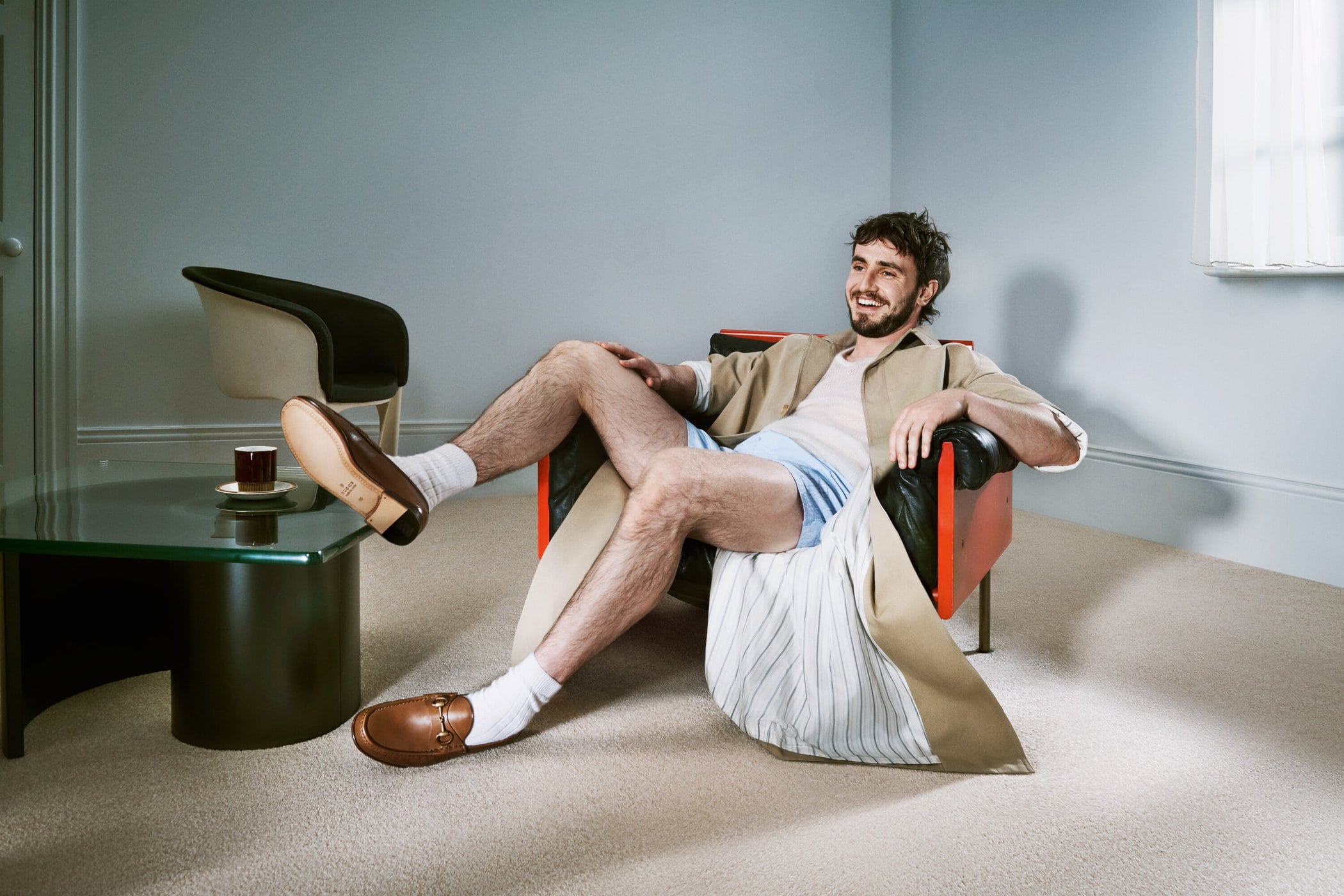 Man in blue shorts and beige coat sits in an armchair in a light gray room. A glass table and chair are in the background.