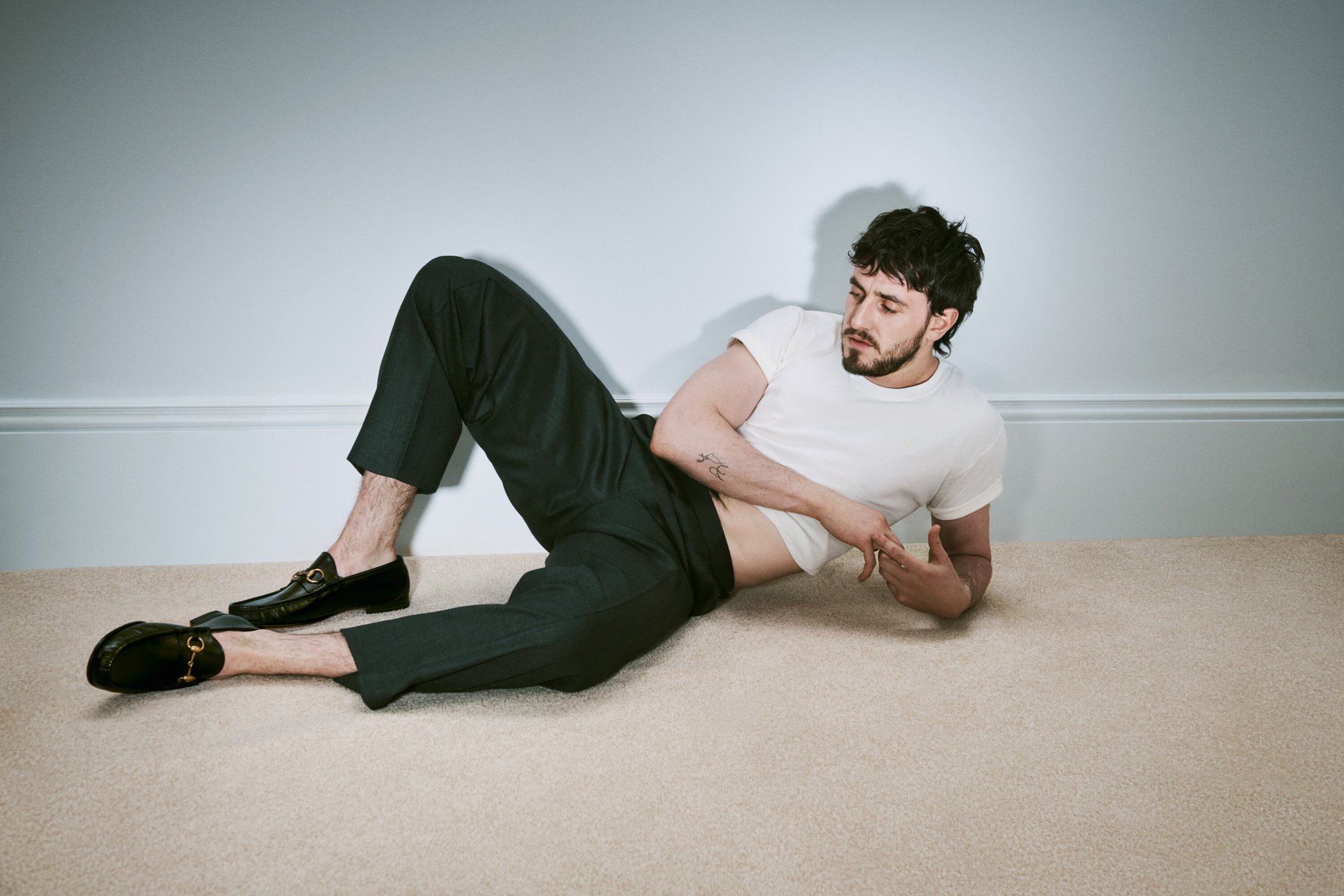 Stylish man in a white t-shirt and dark green pants lies on a beige carpet in front of a light blue wall, posing in black loafers.