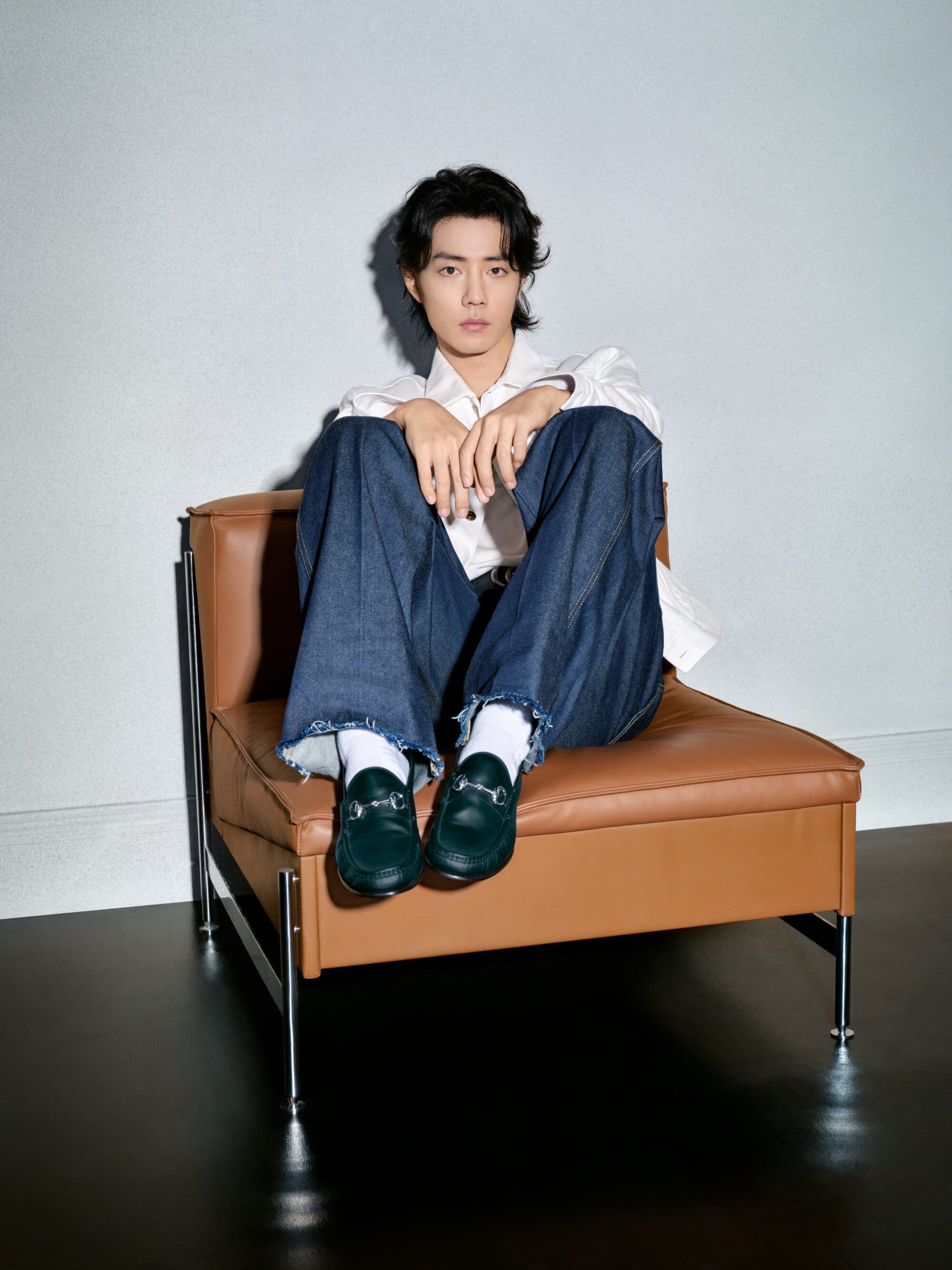 Portrait of an adult male seated in a brown leather chair wearing a white shirt, blue jeans, green loafers, and white socks in a studio.
