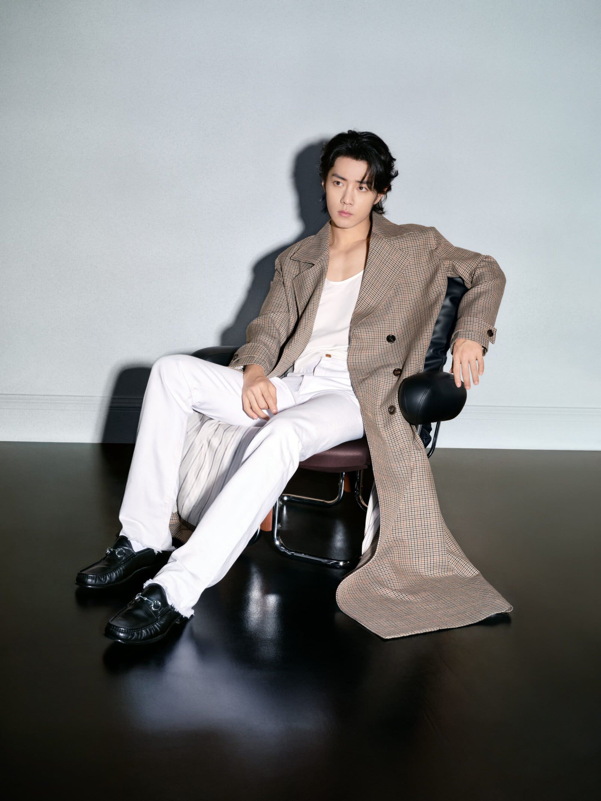 A stylish young man with dark hair and a tan coat poses while sitting in a chair, set against a gradient gray studio backdrop.