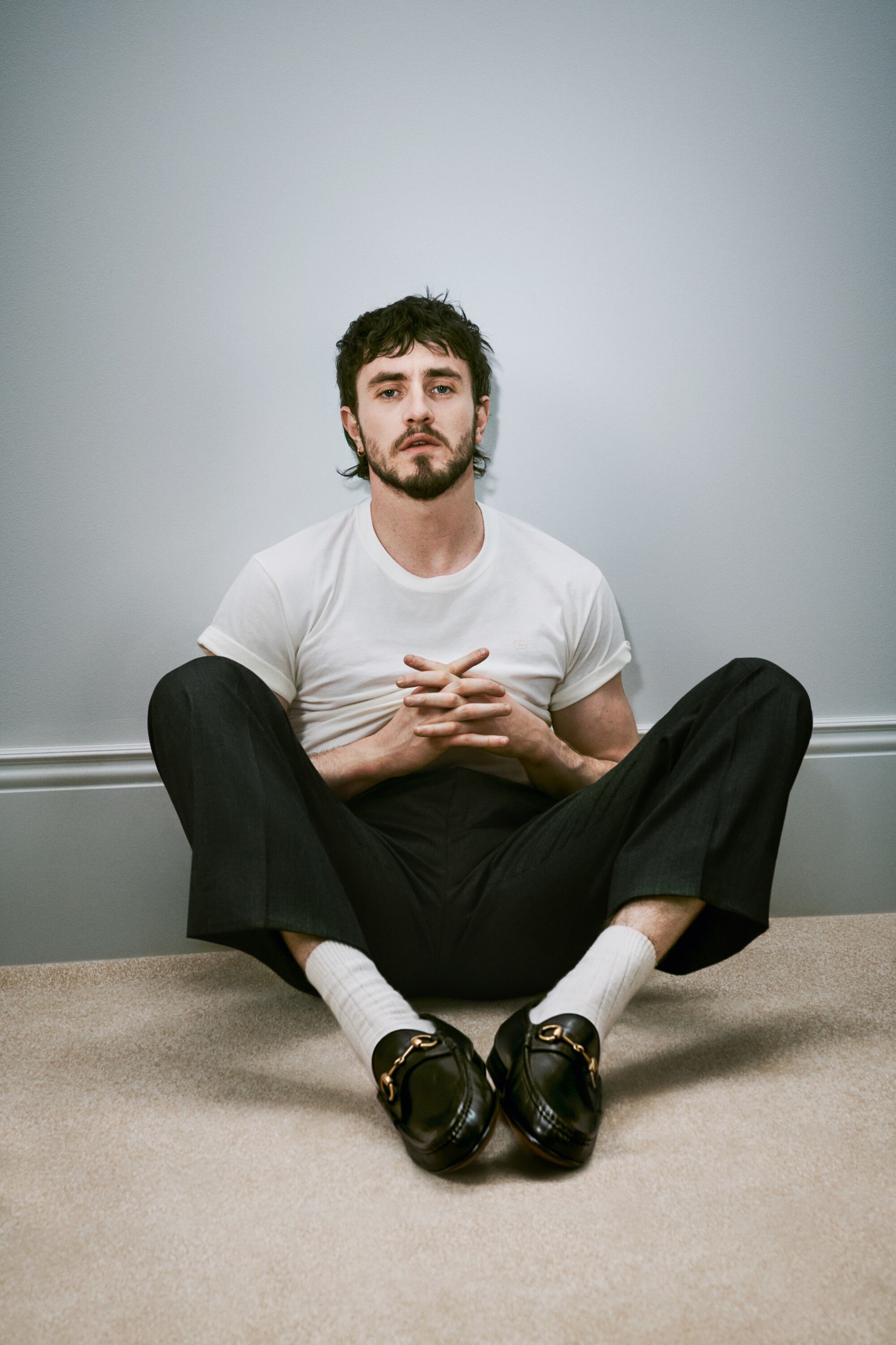 Indoor portrait of a handsome adult man with dark hair and a beard. He's sitting on the floor wearing casual clothes and loafers.