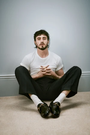 Indoor portrait of a handsome adult man with dark hair and a beard. He's sitting on the floor wearing casual clothes and loafers.