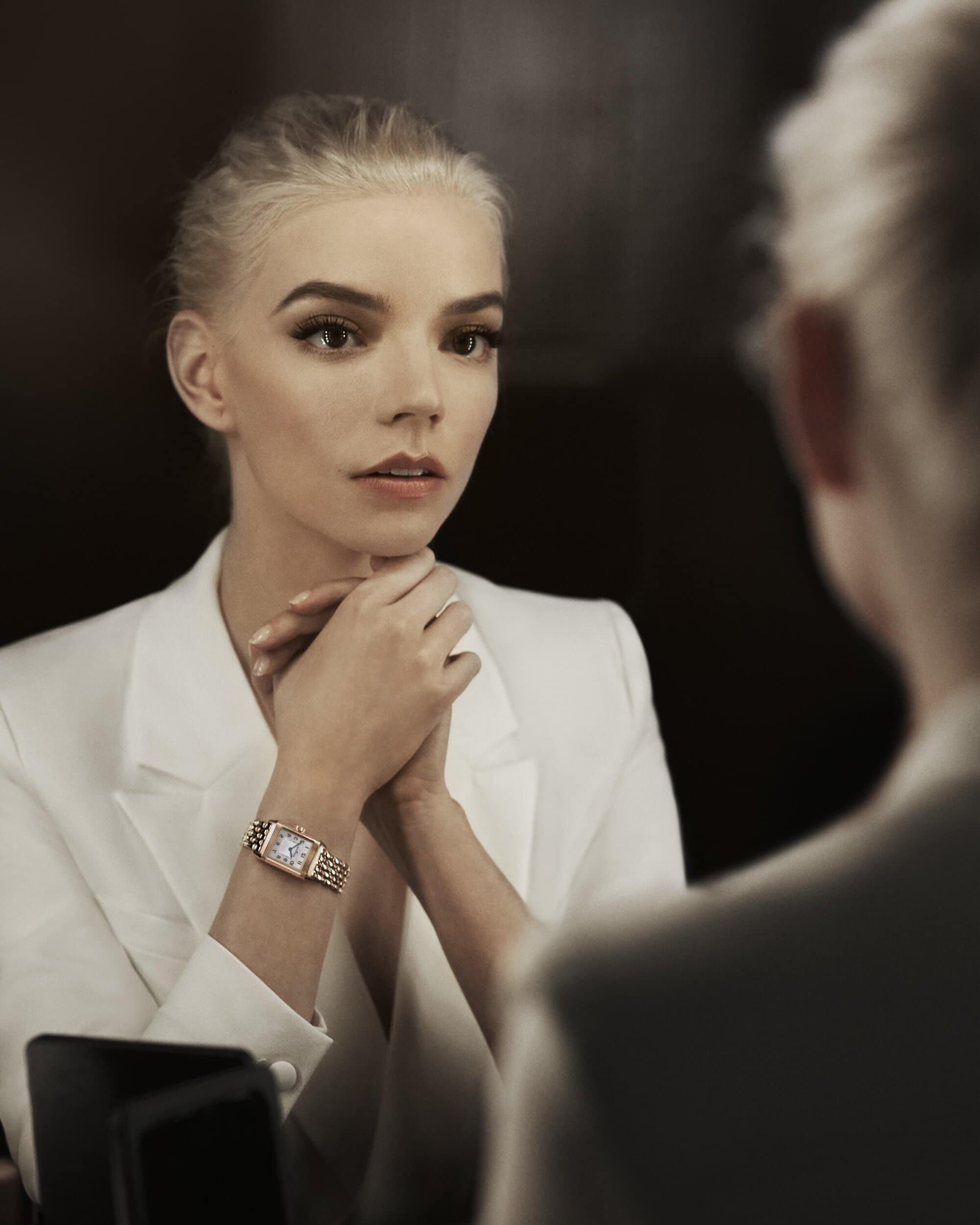 Anya Taylor-Joy gazes in a mirror, wearing an elegant white blazer and a gold watch. Soft lighting highlights her features.