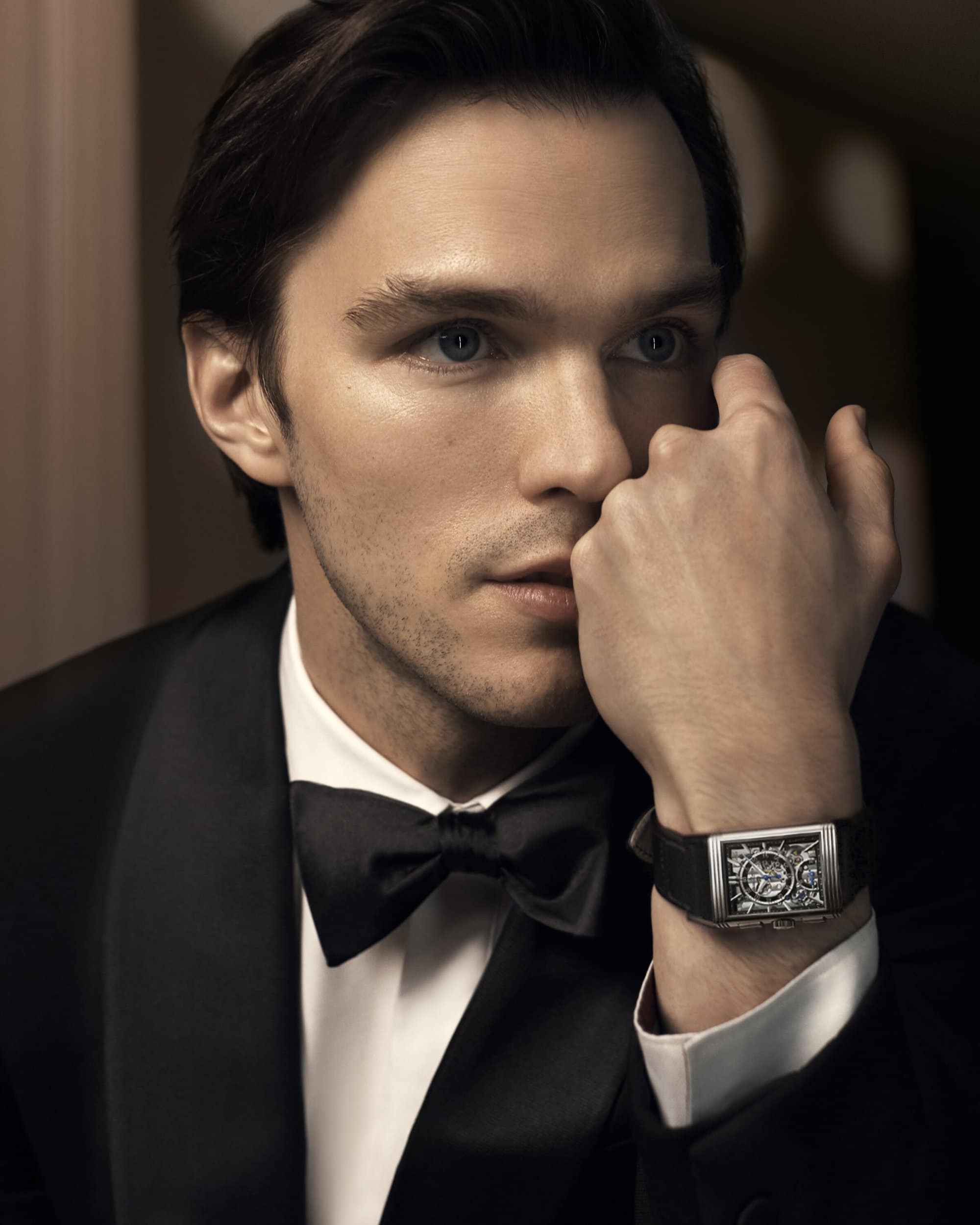 Close-up of adult male in tuxedo and bow tie, wearing a rectangular wristwatch with a black leather strap, looking elegant in studio lighting.