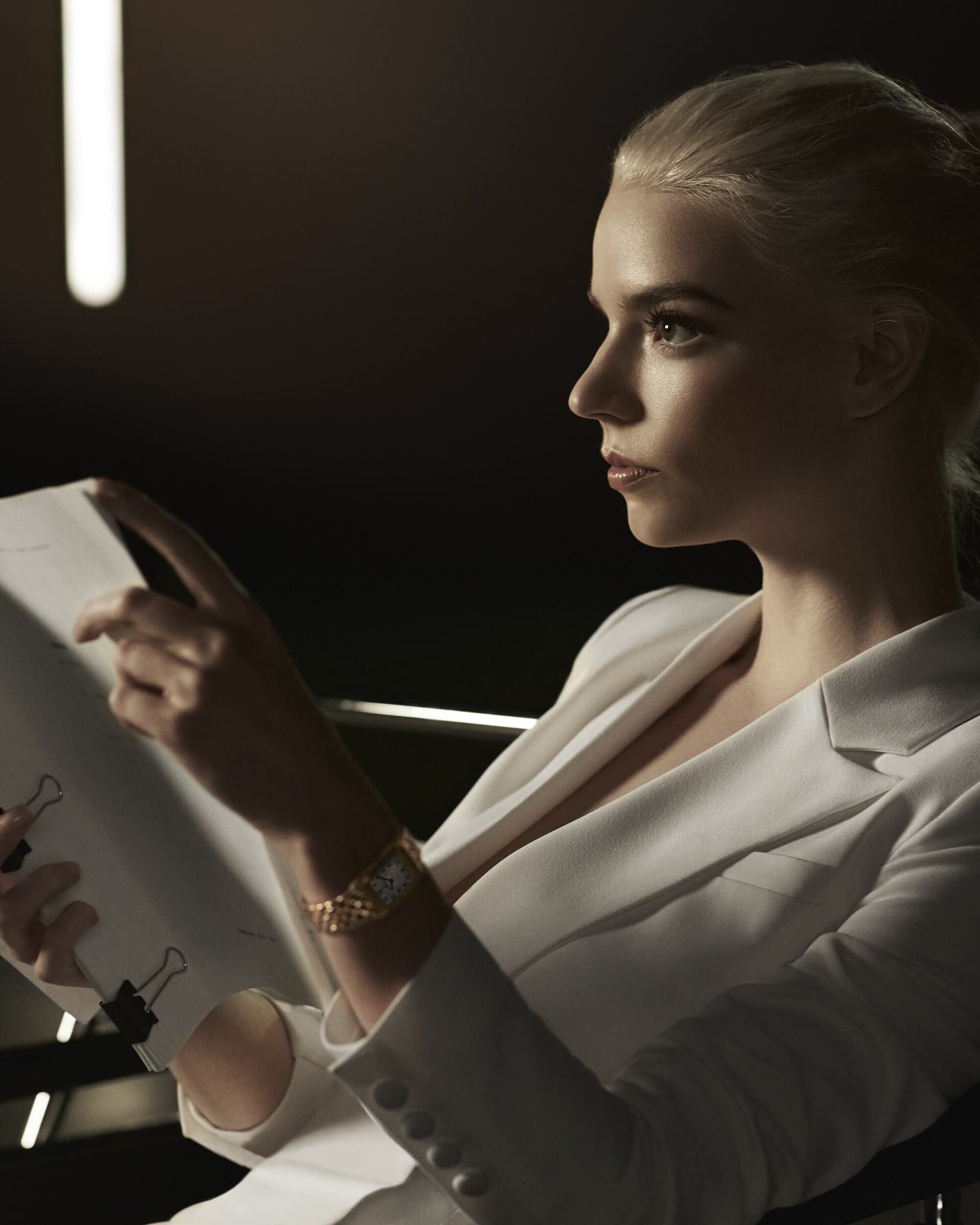 Adult woman in white suit jacket poses indoors with paper document. Dark background, shallow focus, elegant gold wrist watch, soft light.