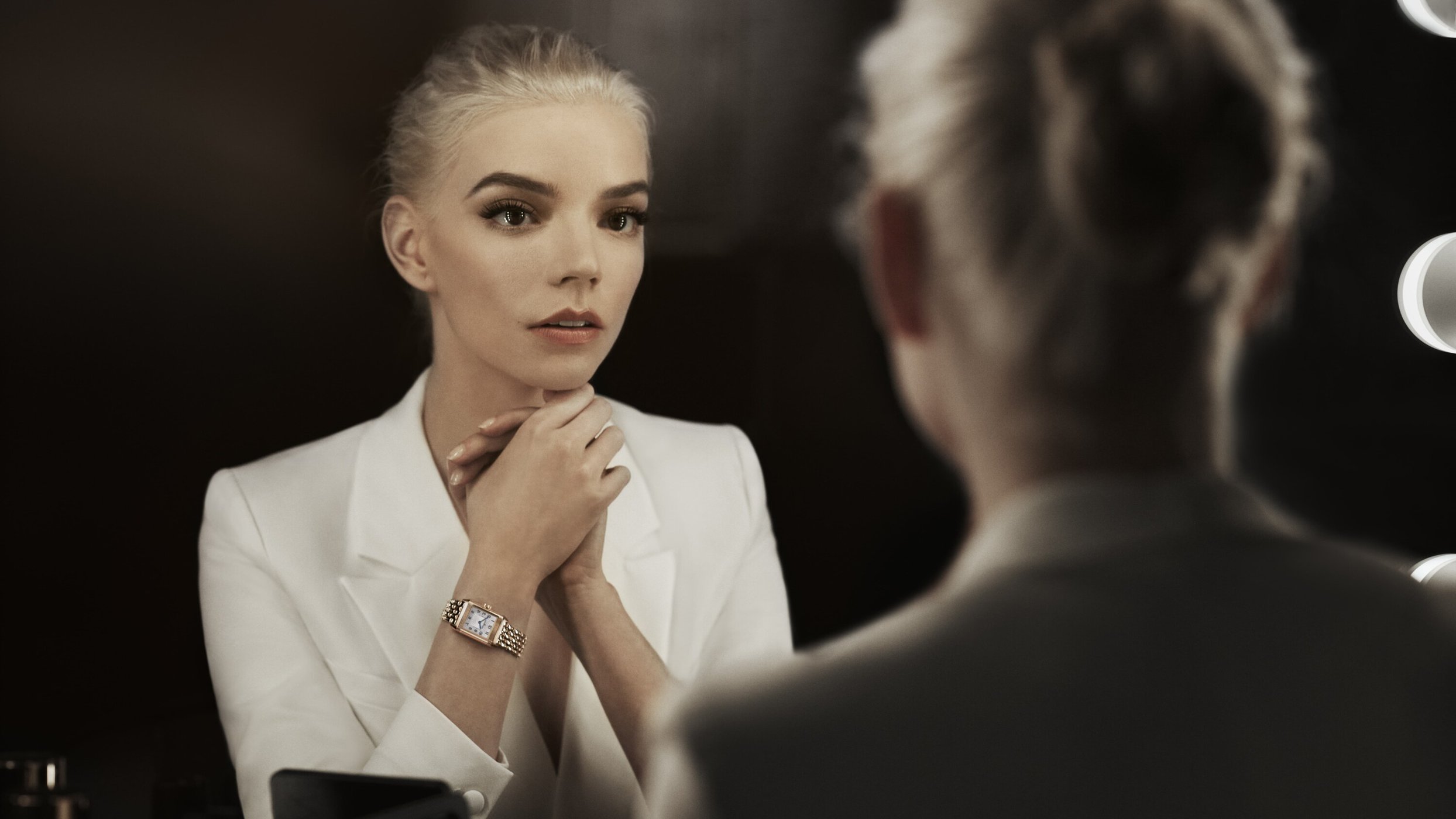 Anya Taylor-Joy in white blazer gazes into a mirror, highlighting a gold wristwatch, set against a dark background with bulb lighting.
