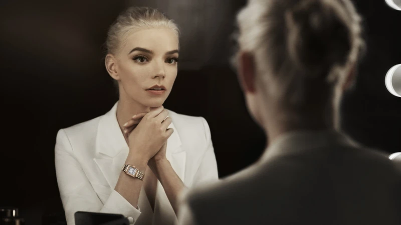 Anya Taylor-Joy in white blazer gazes into a mirror, highlighting a gold wristwatch, set against a dark background with bulb lighting.