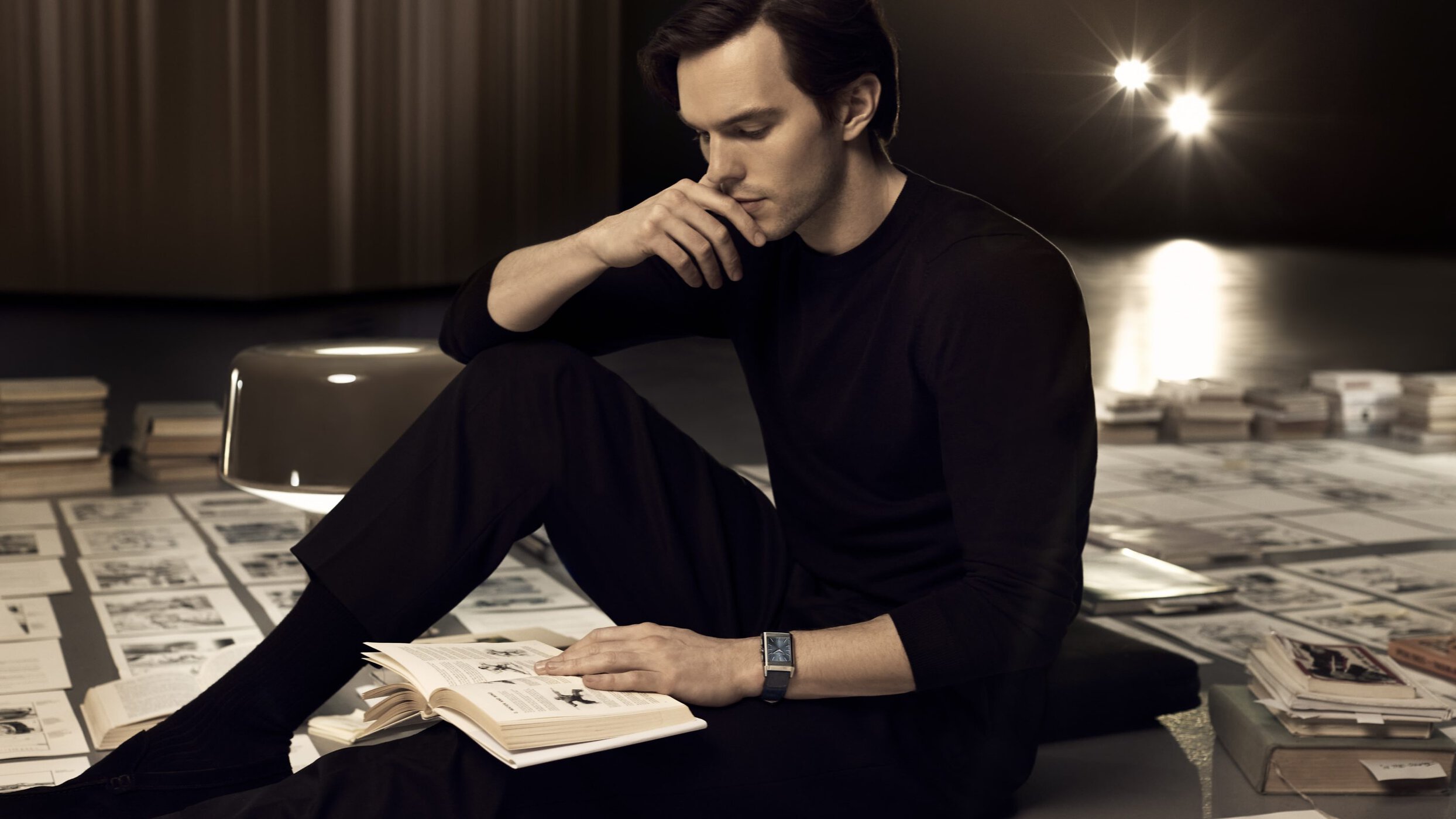 Man in black attire sitting on the floor reading a book surrounded by papers and books in a dimly lit indoor setting.
