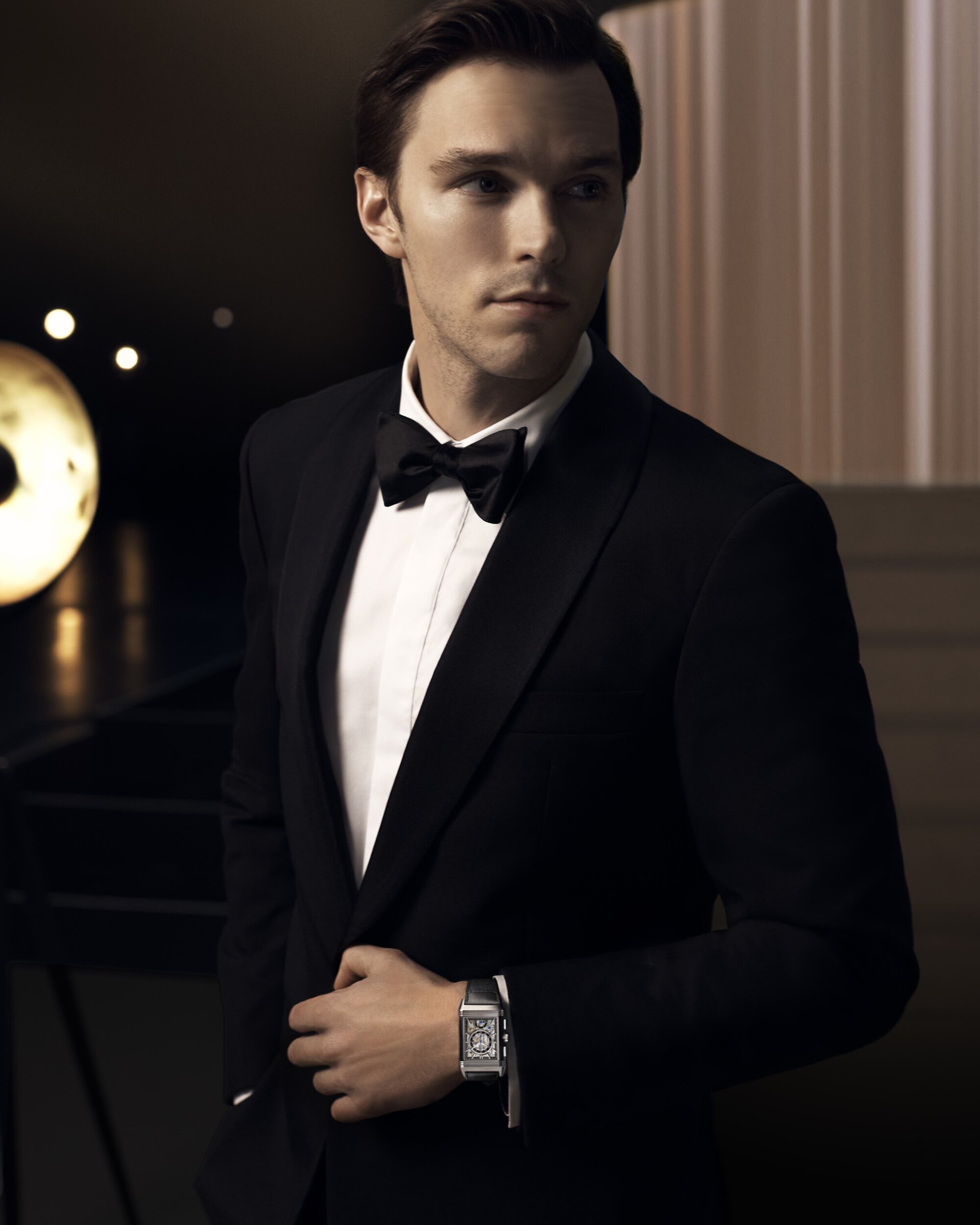 Elegant portrait of a brunette adult male in a classic black tuxedo with a bow tie, complemented by a wristwatch in a dark, moody studio.