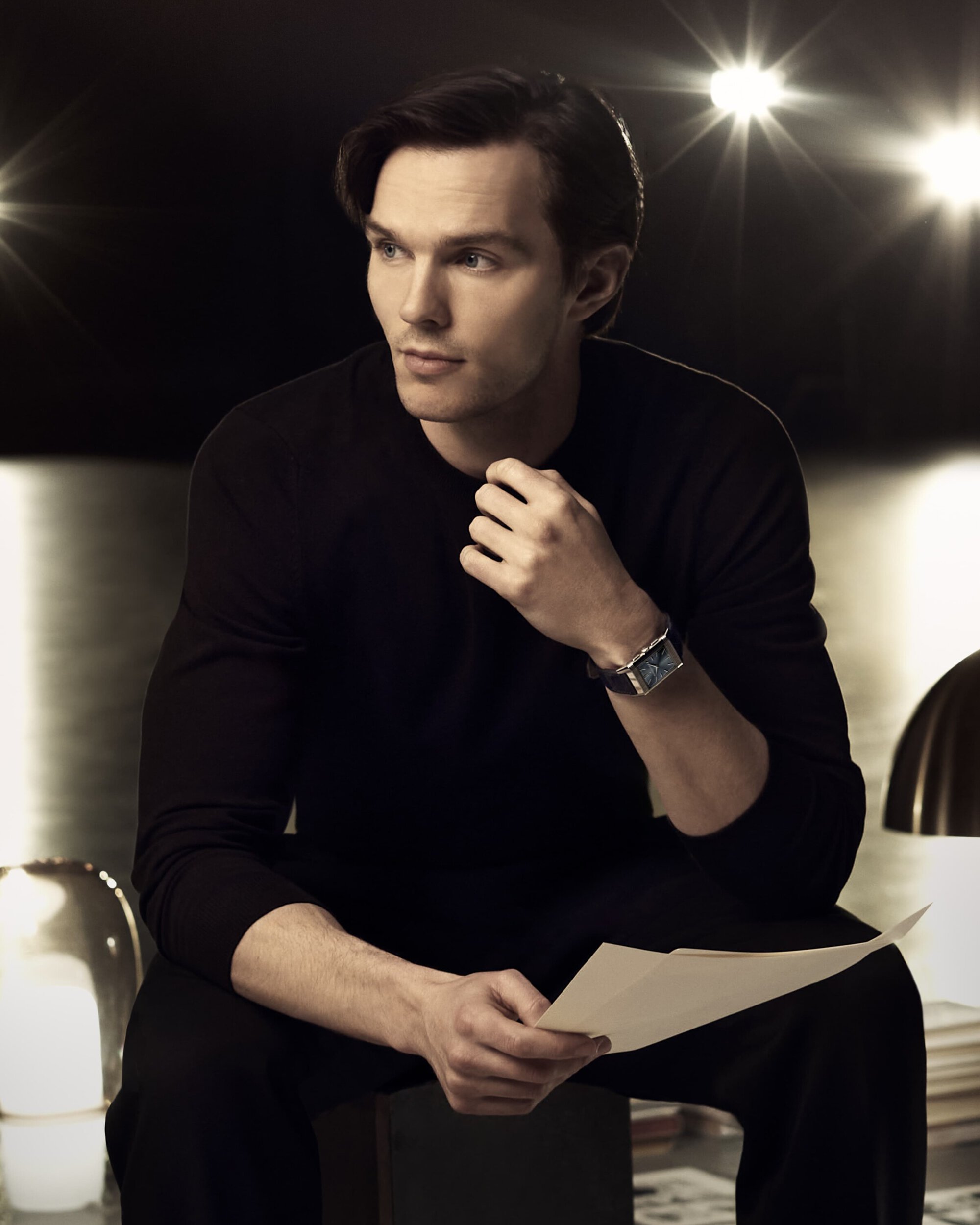Portrait of an adult man in a black shirt sitting, holding a paper, and wearing a watch, with a blurred spotlight background.