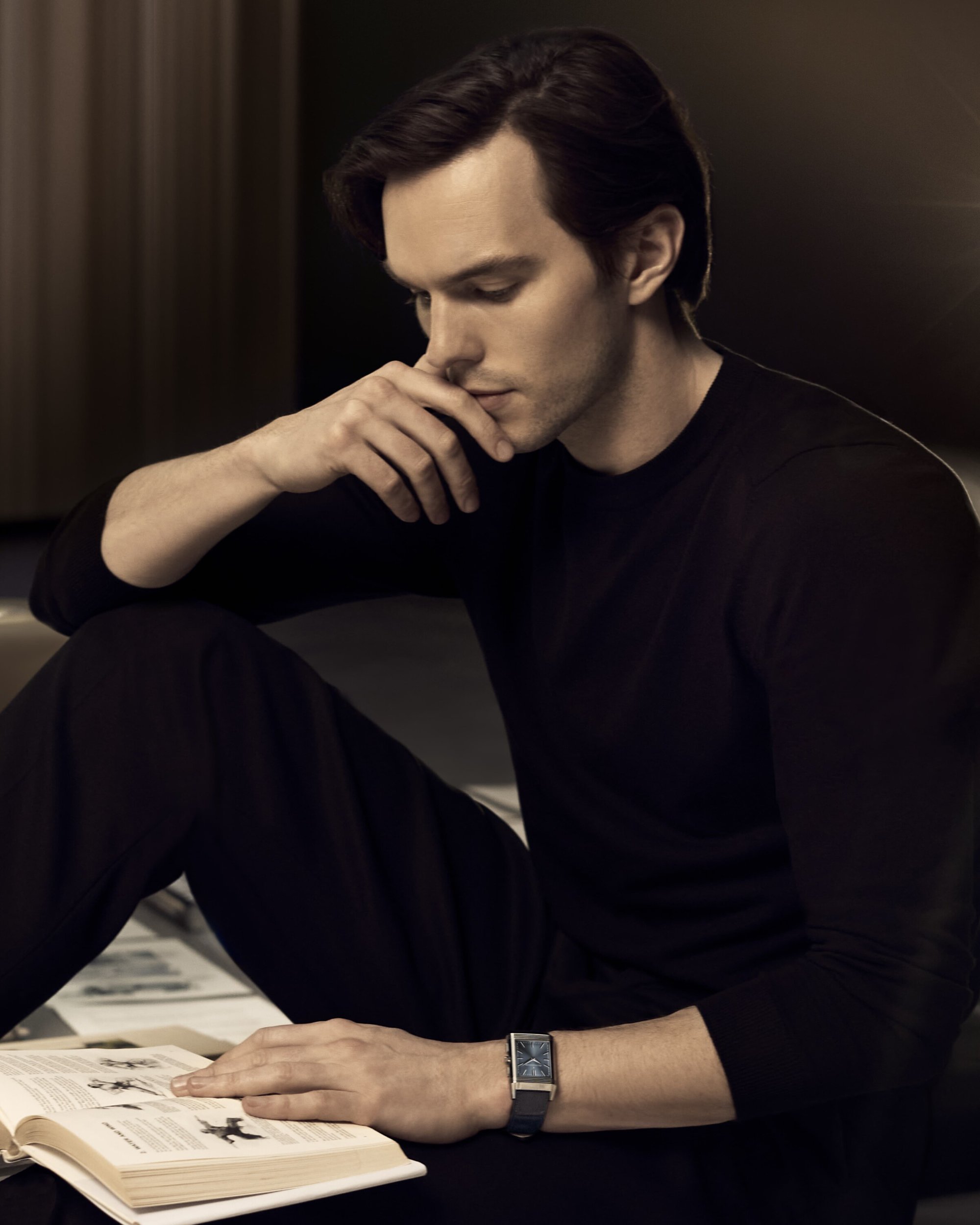 Portrait of a light-skinned man in a black long-sleeved shirt sitting with a book in his lap, looking down indoors.