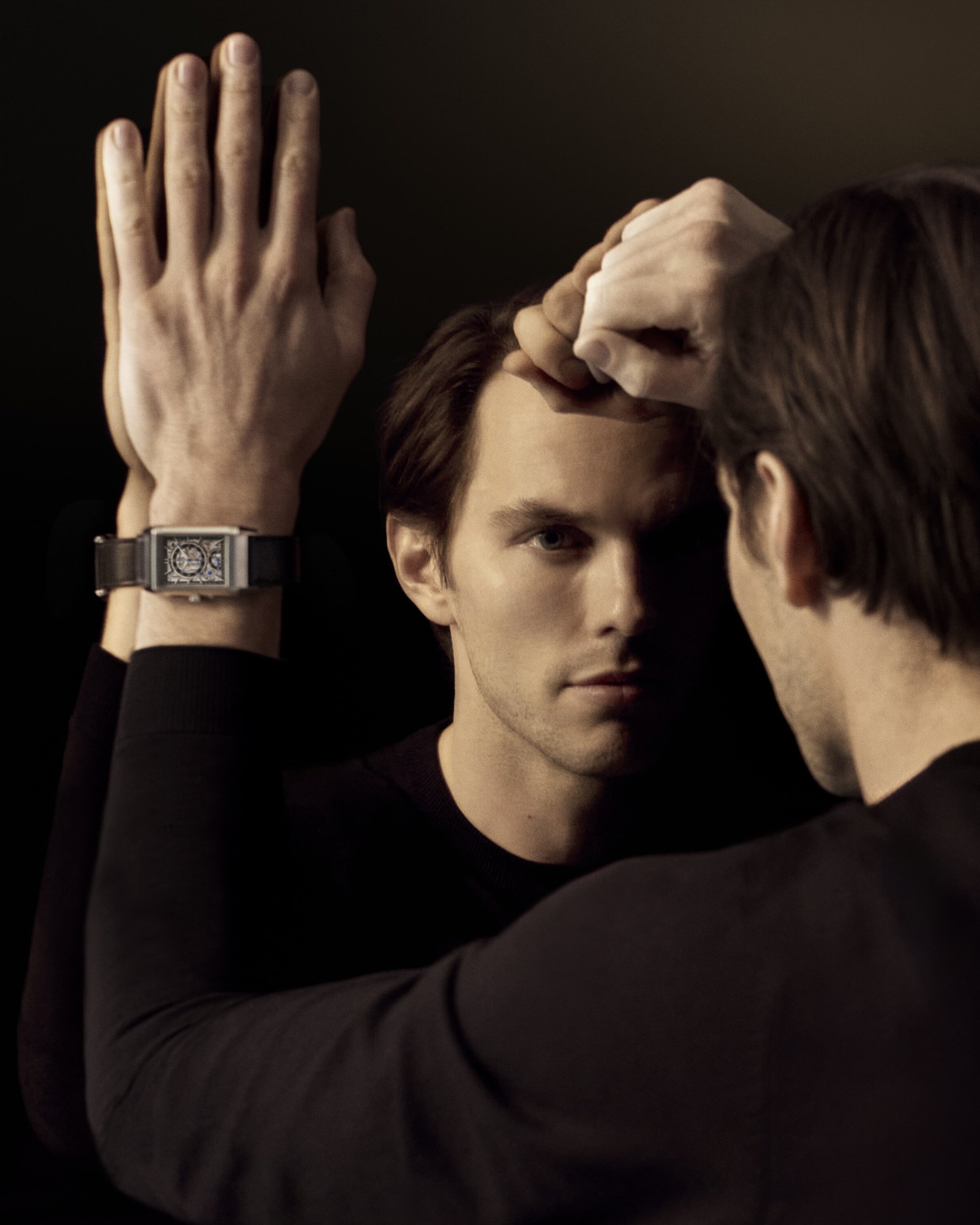 A man in black with a wristwatch poses against a dark background, his reflection visible in the mirror he's leaning against.