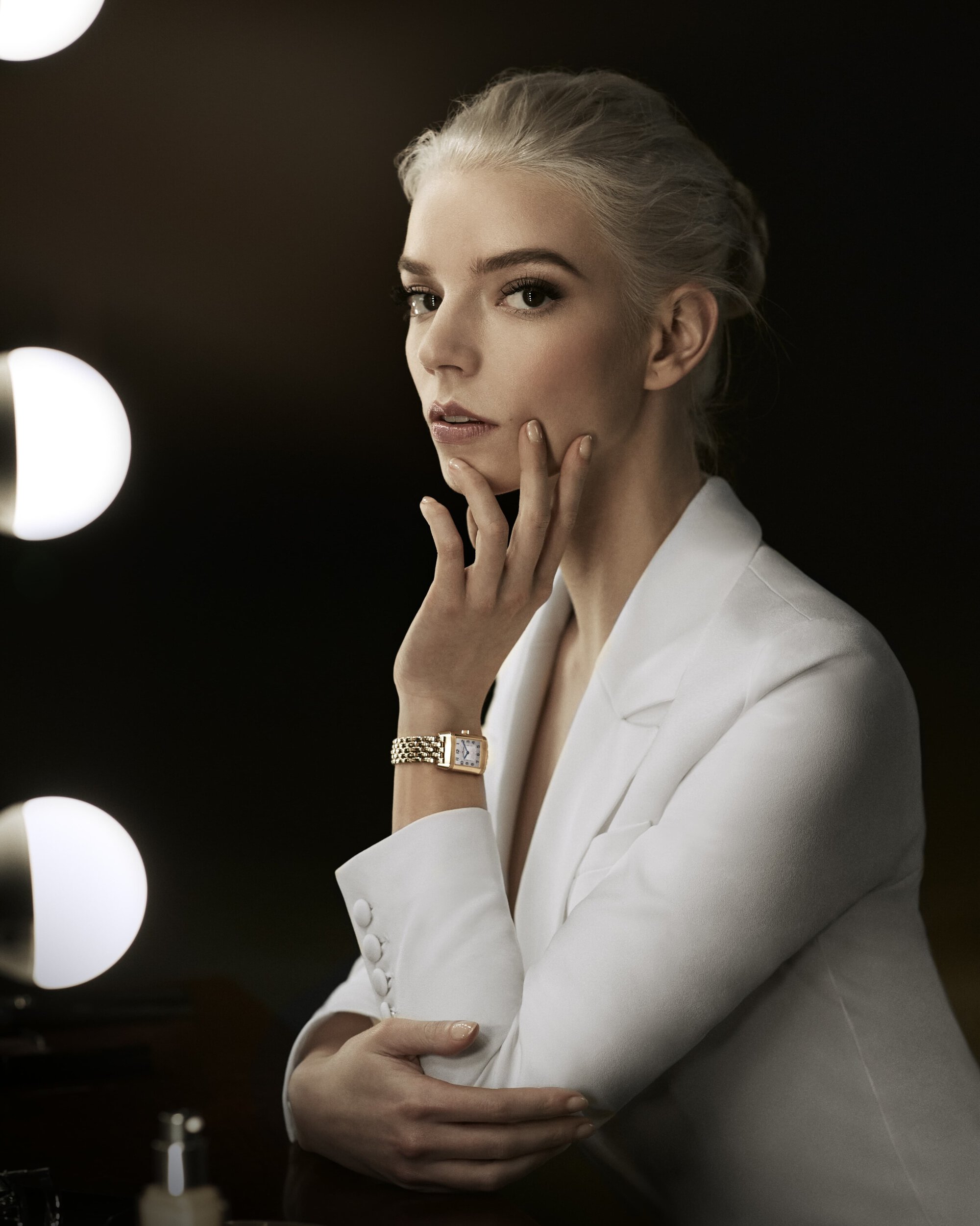 Elegant woman with platinum blonde hair and white suit poses in front of a lighted vanity mirror, showcasing a gold watch.