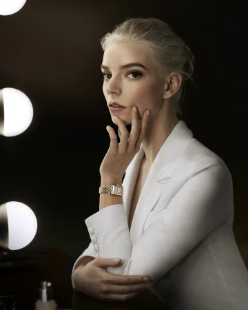 Elegant woman with platinum blonde hair and white suit poses in front of a lighted vanity mirror, showcasing a gold watch.