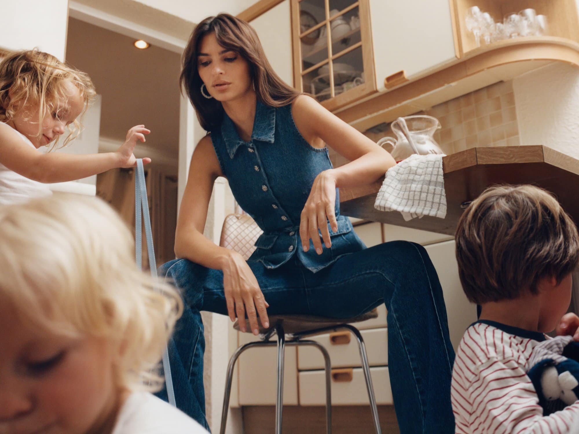 Emily Ratajkowski sits on a stool in a denim vest and jeans in a kitchen with three children playing and looking at her.
