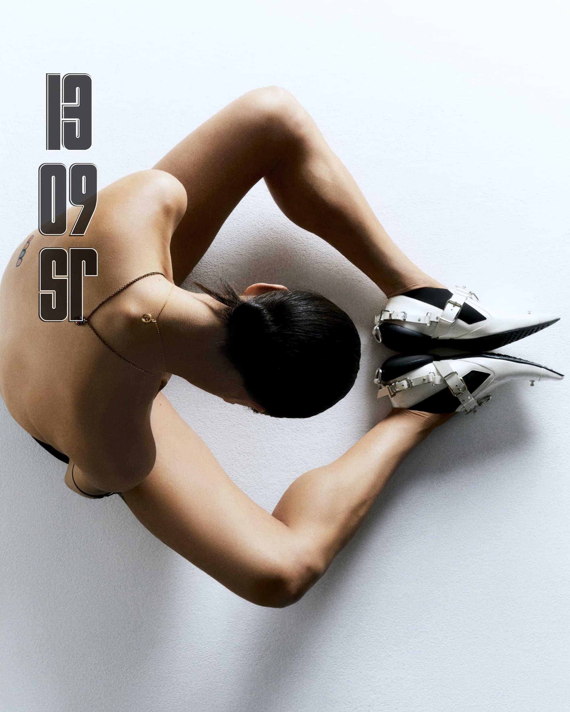 Overhead shot of a person lying on a white surface wearing white and black shoes with silver buckles, and the numbers '13 09 SR'.