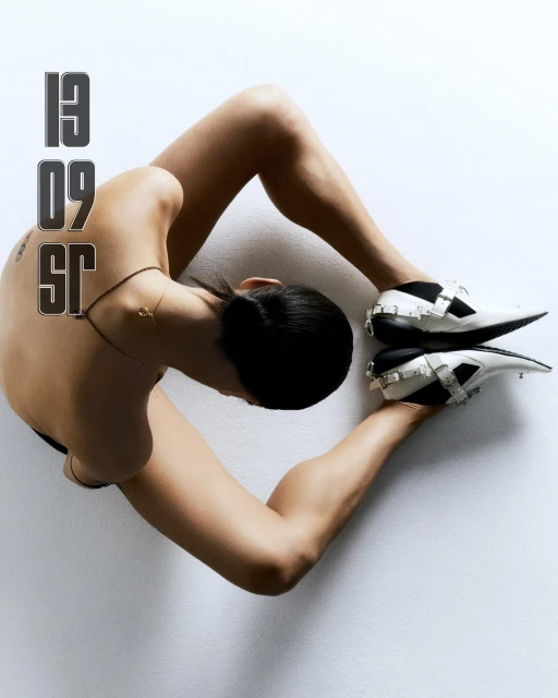 Overhead shot of a person lying on a white surface wearing white and black shoes with silver buckles, and the numbers '13 09 SR'.