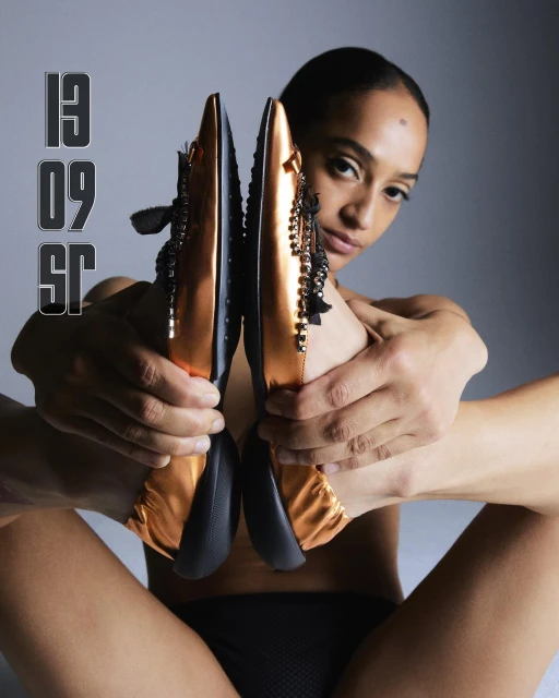 Elegant bronze-and-black ballet flats are held by a bare-chested model in an indoor studio setting, against a diffused gradient backdrop.