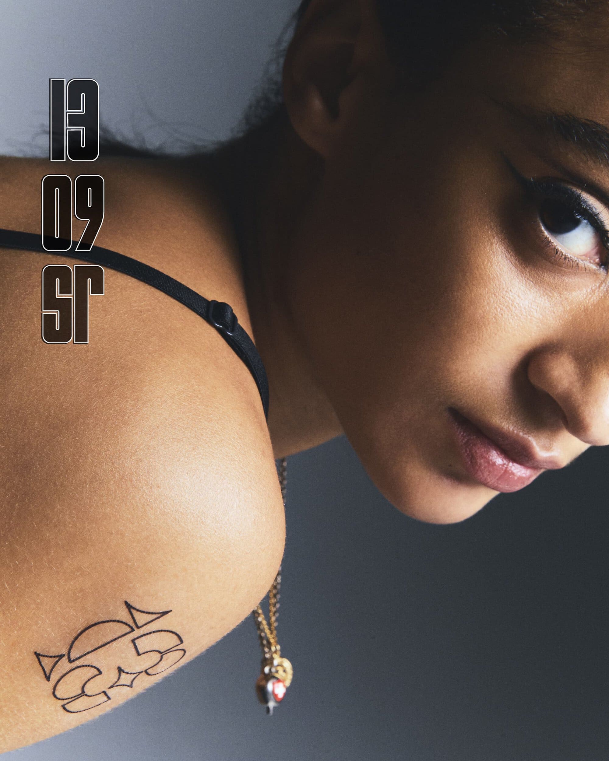 Close-up studio shot of a young adult female with a tattoo, wearing a black top and gold necklace, against a grey background.
