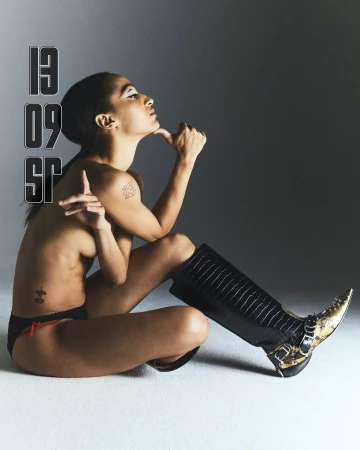 Letizia Galloni seated in profile wearing black knee-high boots and swimwear briefs against a neutral studio backdrop.