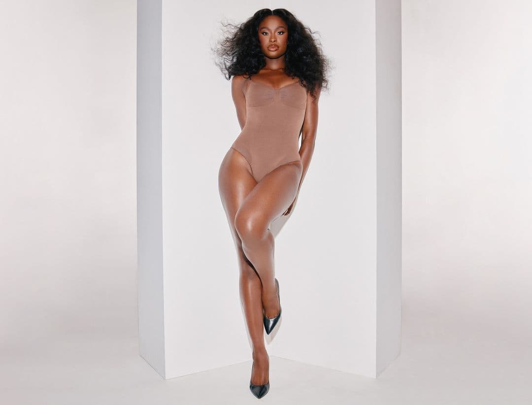 A woman in a tan bodysuit and black high heels poses next to a white corner. Her hair is voluminous, and she looks at the camera.