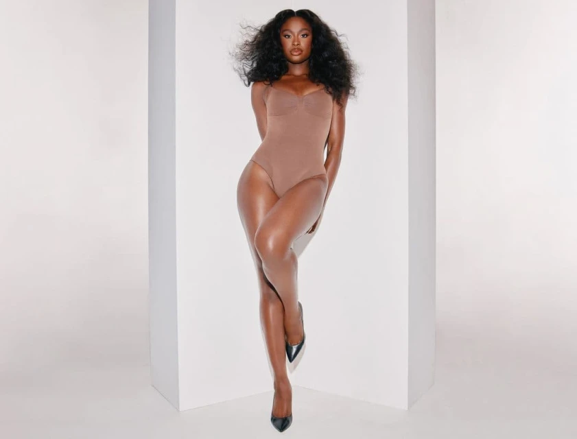 A woman in a tan bodysuit and black high heels poses next to a white corner. Her hair is voluminous, and she looks at the camera.
