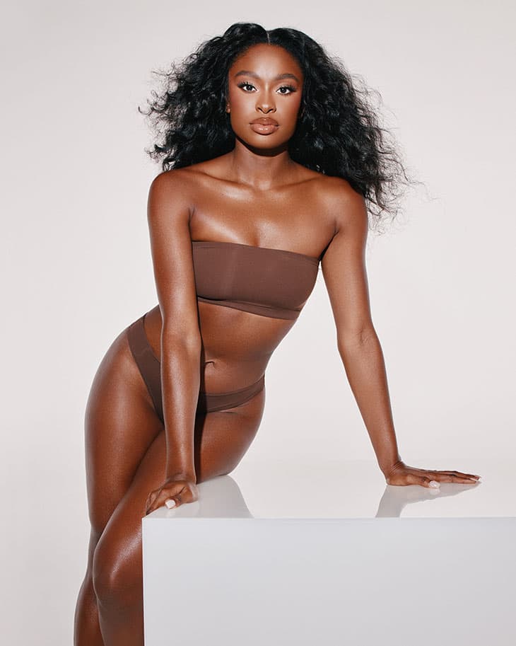 A brunette woman with curly hair poses in a brown bandeau top and bikini bottoms leaning on a white surface in a studio setting.