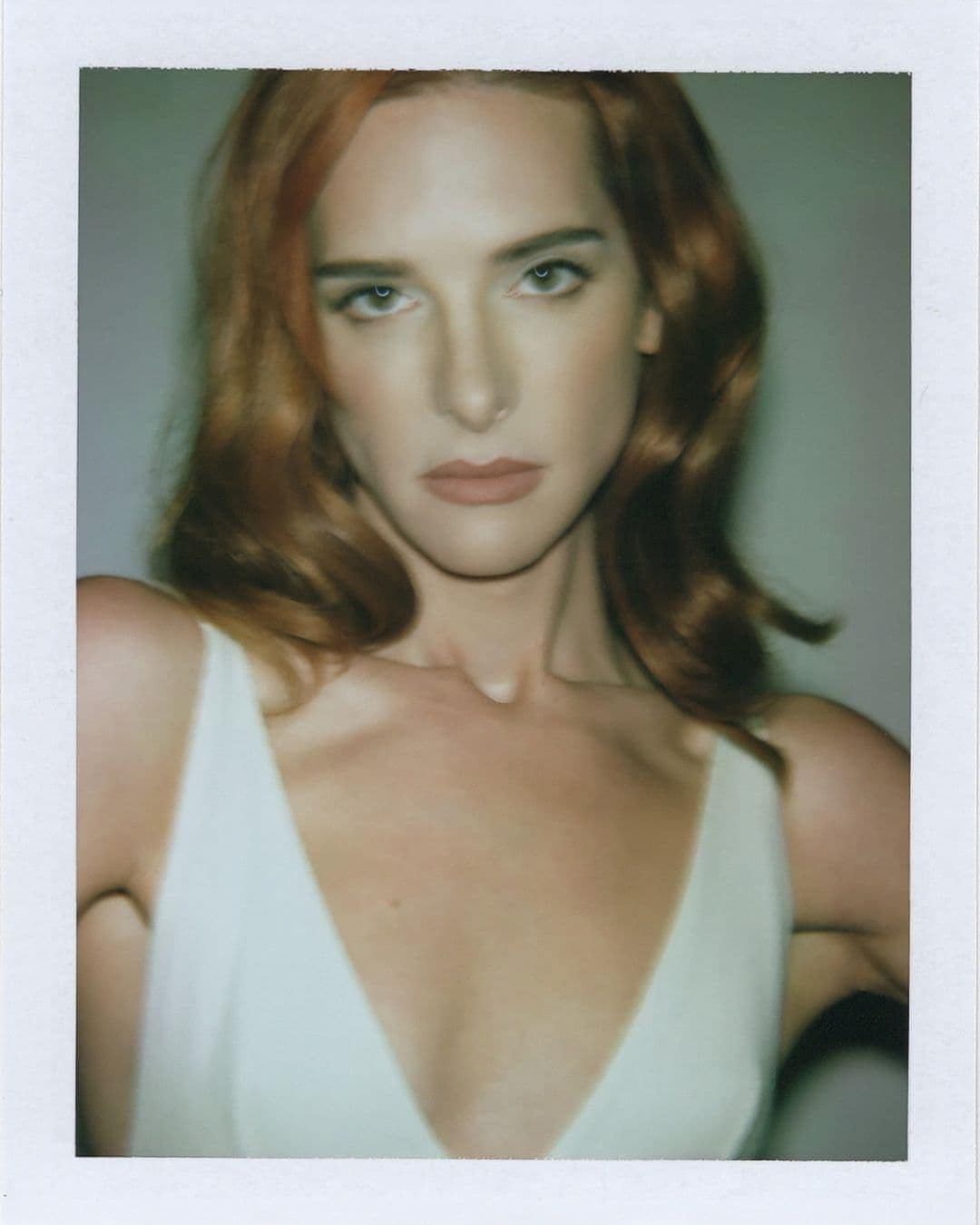 Close-up portrait of an adult woman with auburn hair wearing a white sleeveless dress, looking directly at the camera in a studio setting.