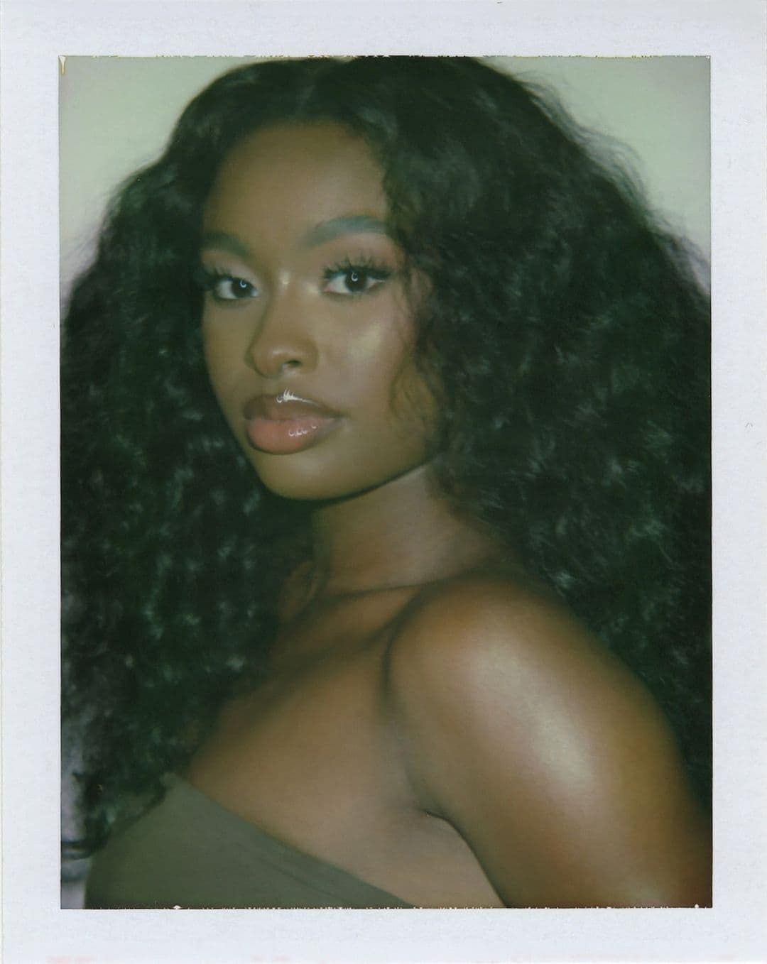 Close-up portrait of a young adult African American woman with long wavy hair, wearing a strapless top, set against a neutral background.