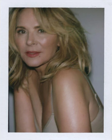 Kim Cattrall in a beige top posing against a plain grey backdrop with a white polaroid border frame