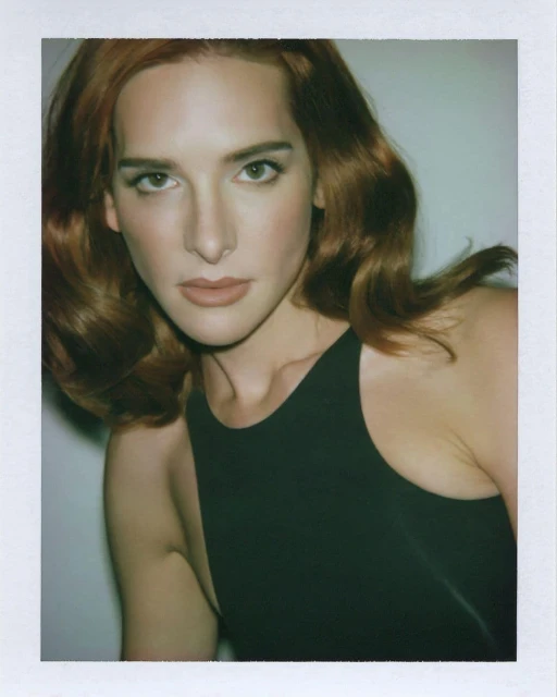 Portrait of a red-haired adult woman in a sleeveless black top against a white background, shot with a close-up framing.