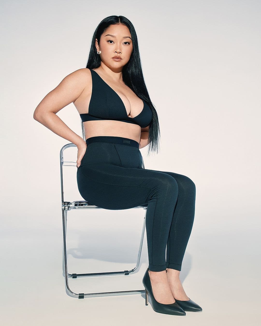 Portrait of an Asian woman in black bra top and leggings seated on a silver chair with a white backdrop in a studio shot.