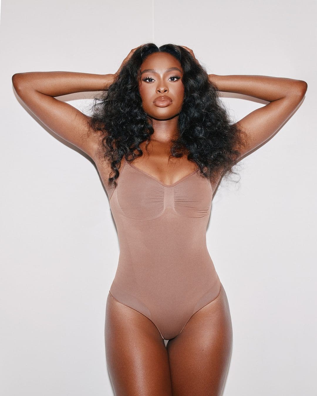 A woman with dark curly hair poses in a nude-colored bodysuit against a white background, her arms up and hands behind her head.