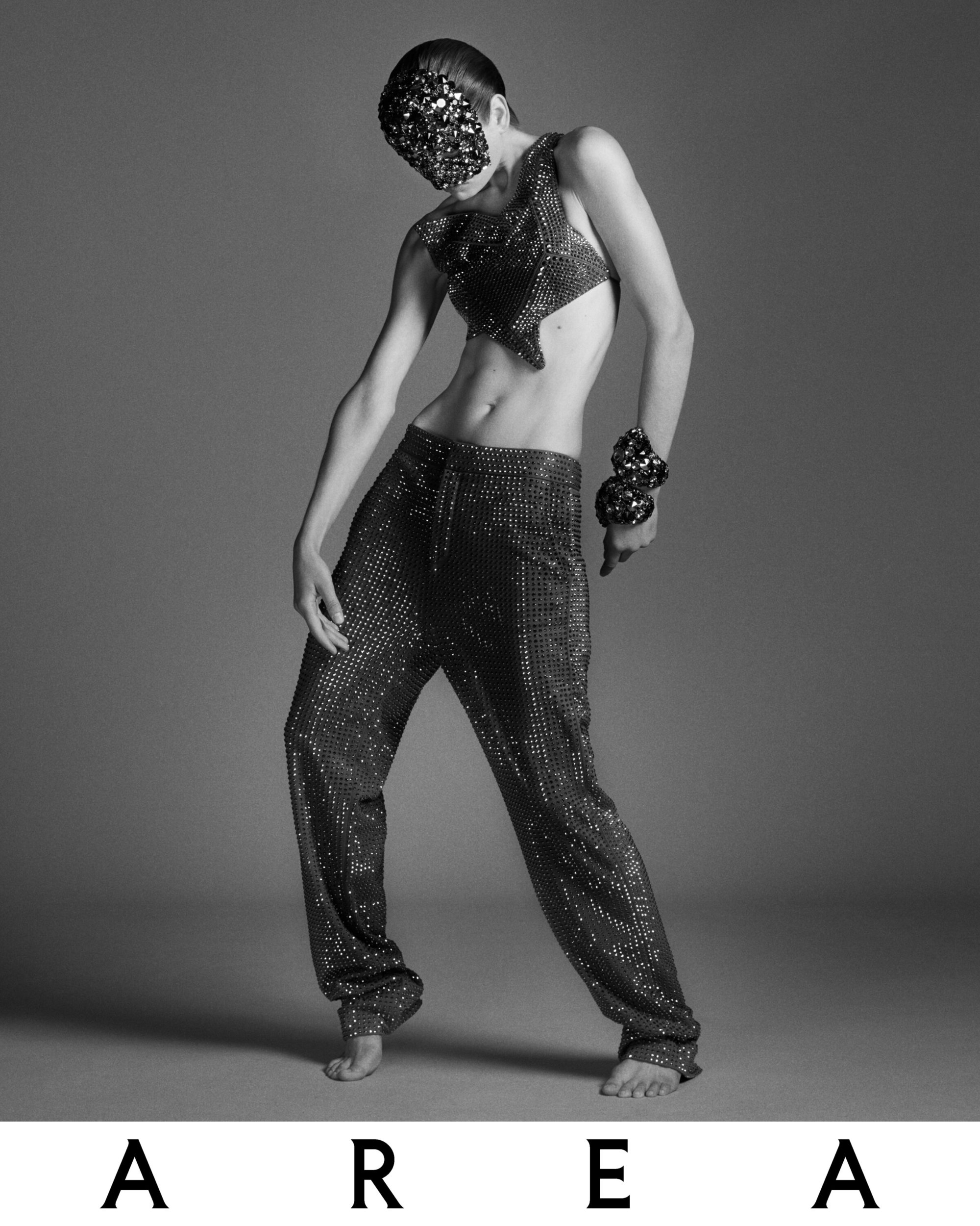 Monochrome fashion shot of a slender model in a sequined top, baggy pants, and a face mask, posing in a studio against a gray background.