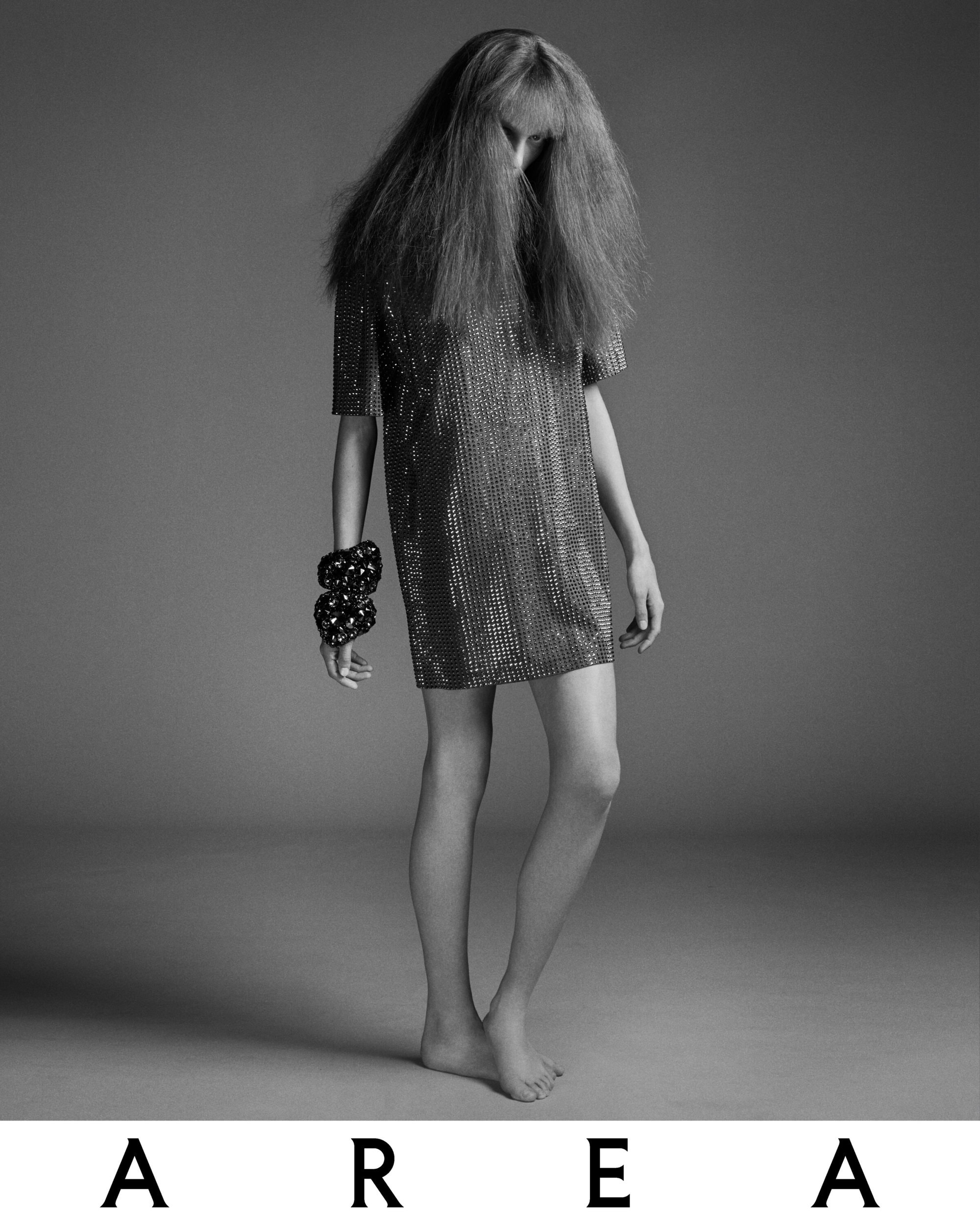 Black and white full shot of a slender model in a metallic mini dress and large bracelets, posed on a seamless gray background.