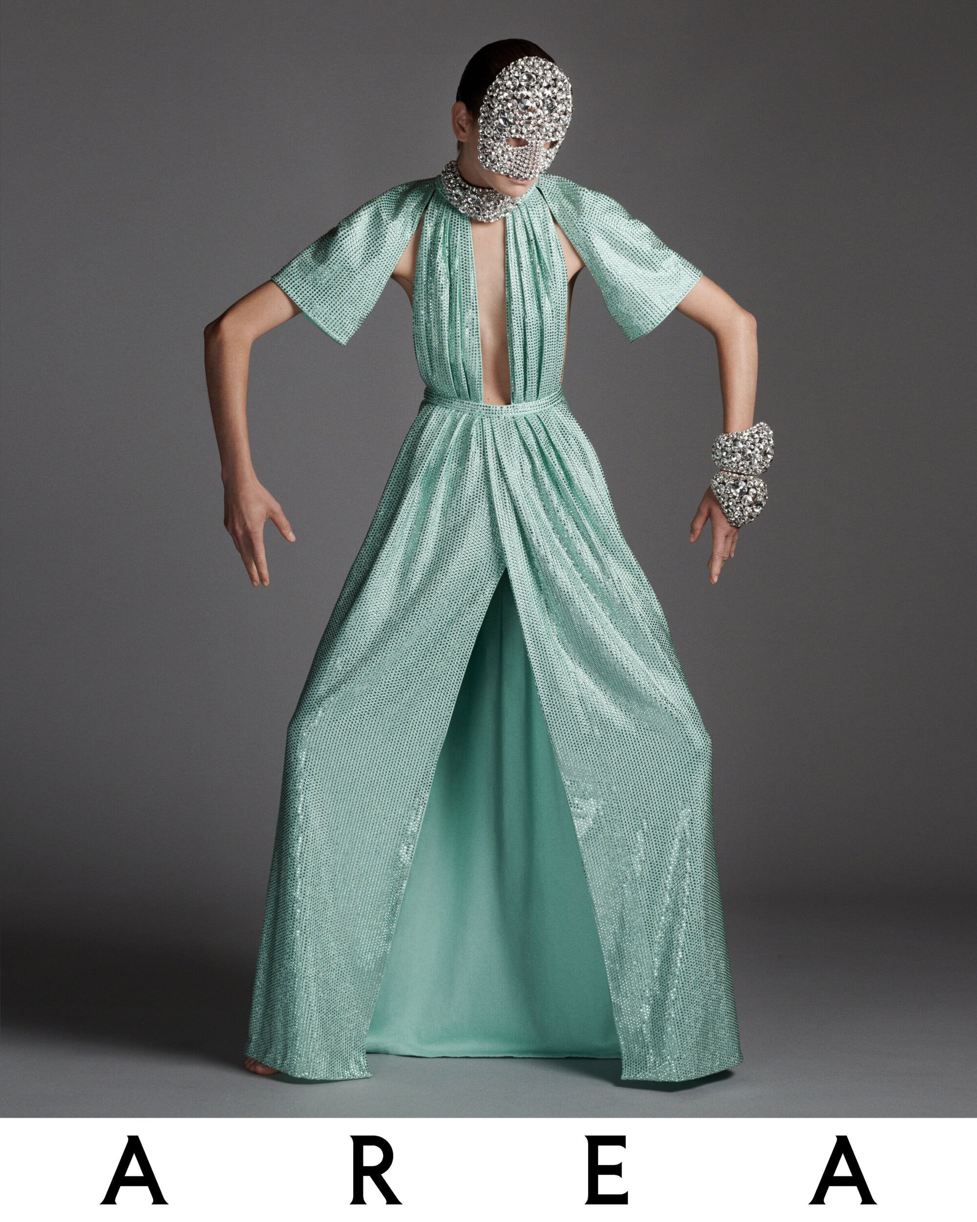 A fashion model poses in a shiny, light-green gown with a crystal mask and matching crystal bracelets against a neutral gray background.