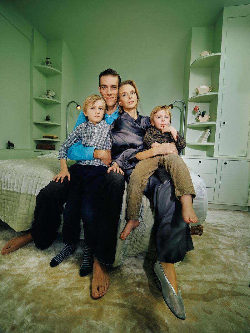 Family portrait indoors: A man and woman sit on a bed with two young children in a light green room with built-in shelves and cabinets.