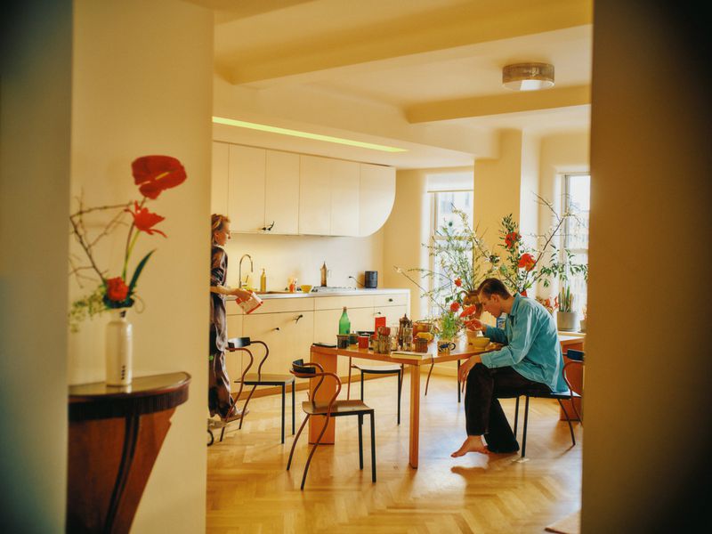 Indoor scene of a man sitting at a table with flowers and food while a woman stands near kitchen counters.